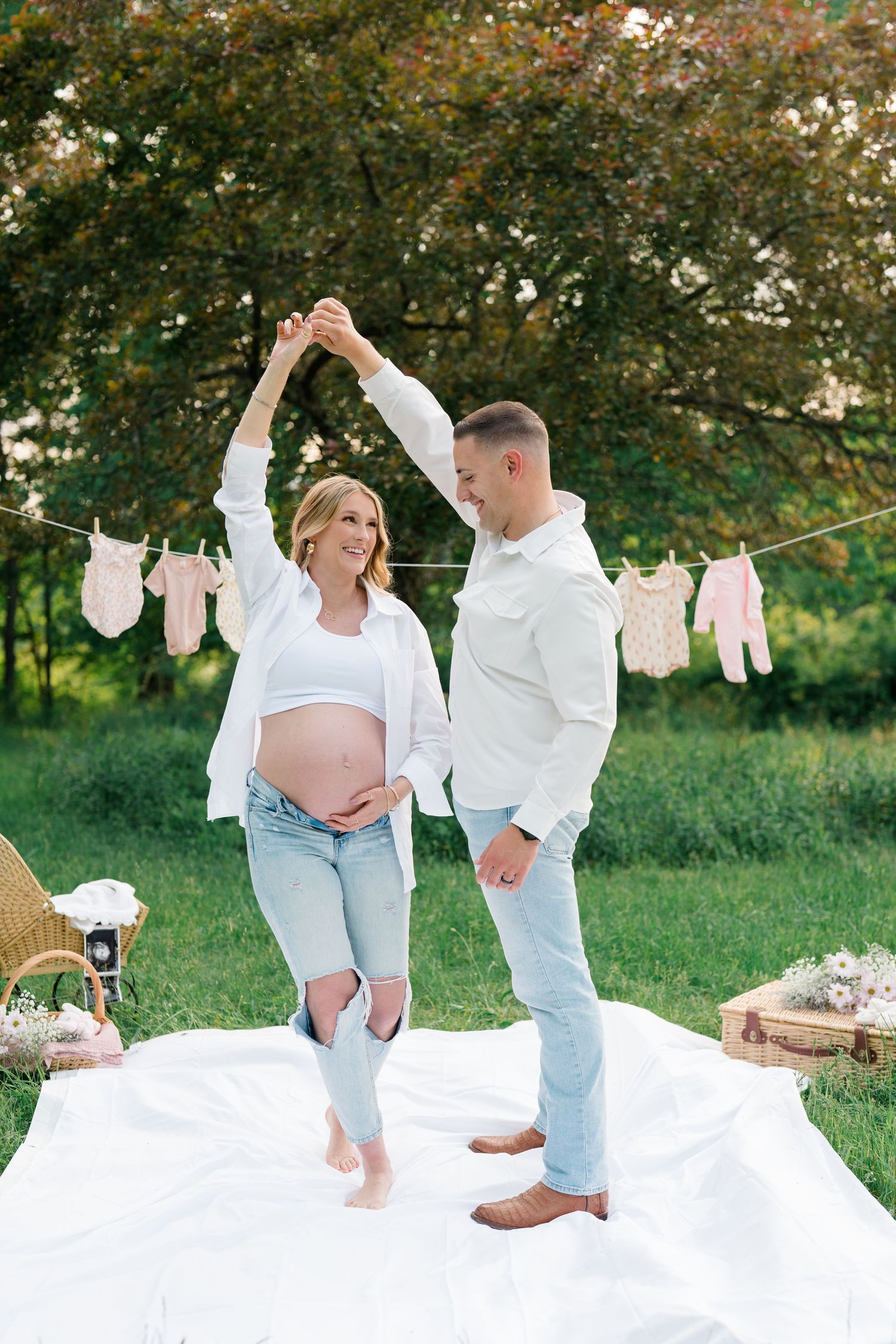 Dancing maternity photoshoot on white blanket with baby clothes hanging on clothesline in garden setting.