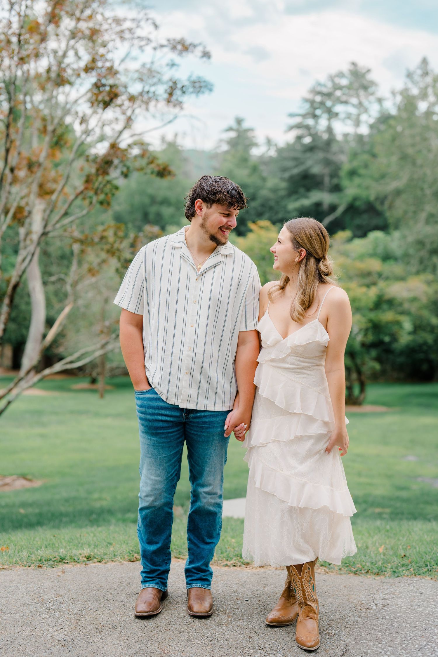 Couple holding hands and smiling at each other in a lush green park, woman in white dress and boots, man in striped shirt.