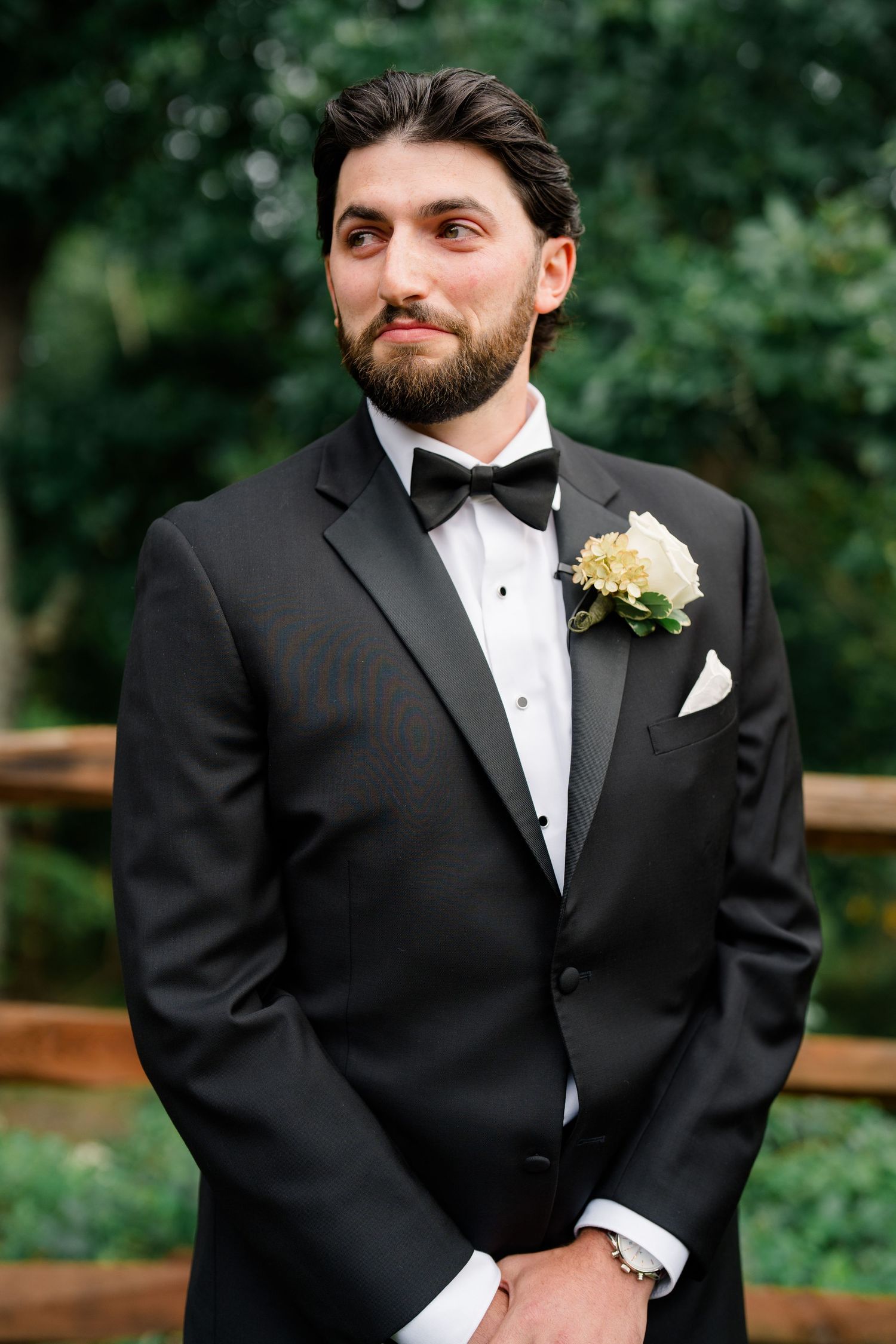 Groom in black tuxedo with bow tie and white floral boutonniere posing outdoors at wedding.