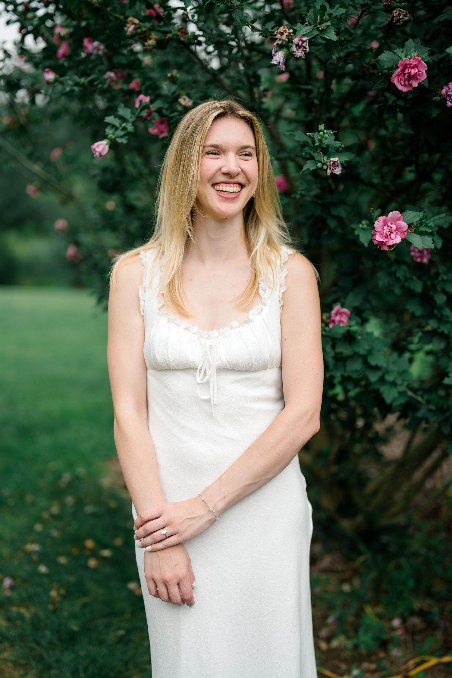 Smiling blonde woman in white lace dress standing by pink roses in a lush green garden.