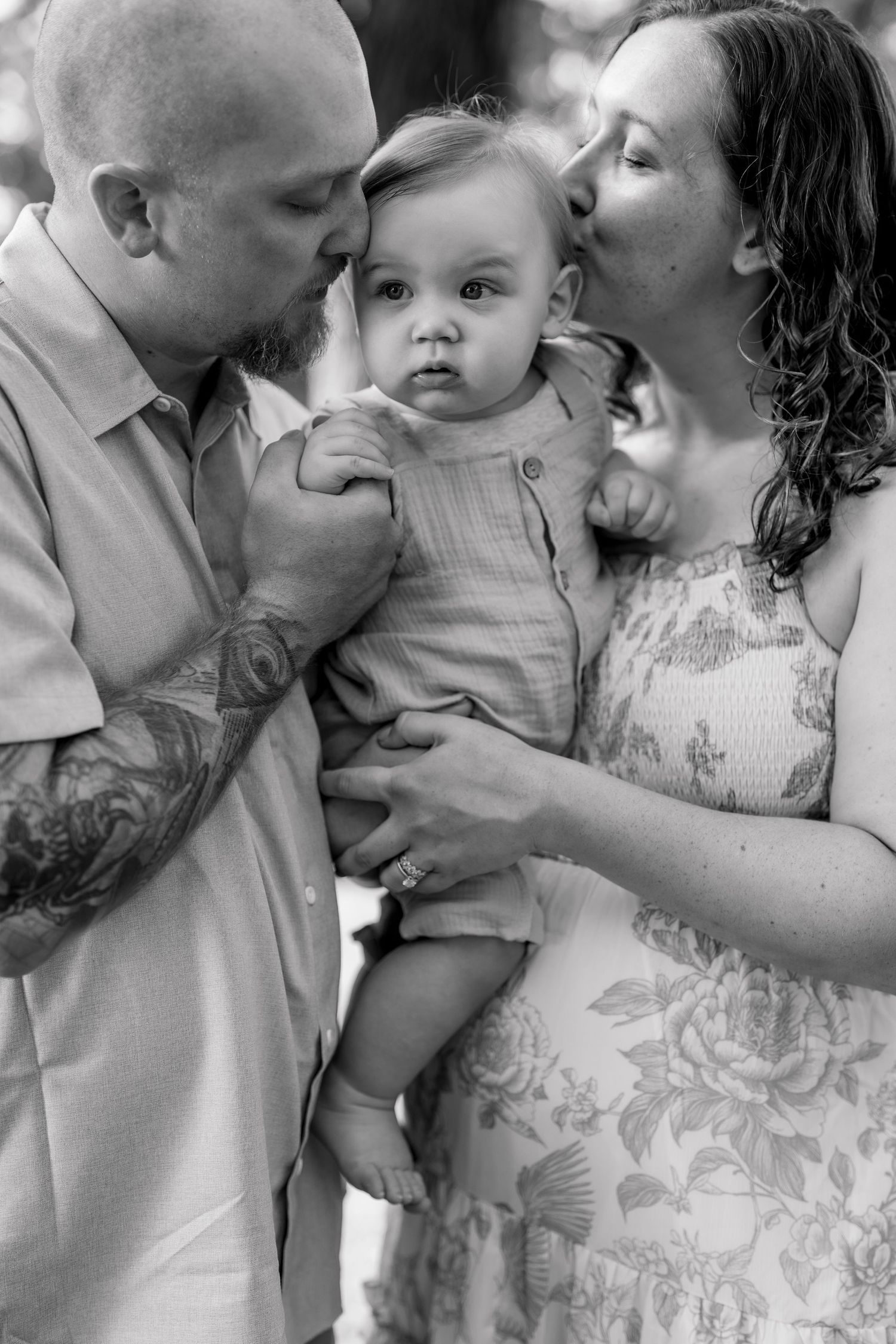 Black and white photo of parents kissing their baby on the cheeks during an outdoor family portrait session.
