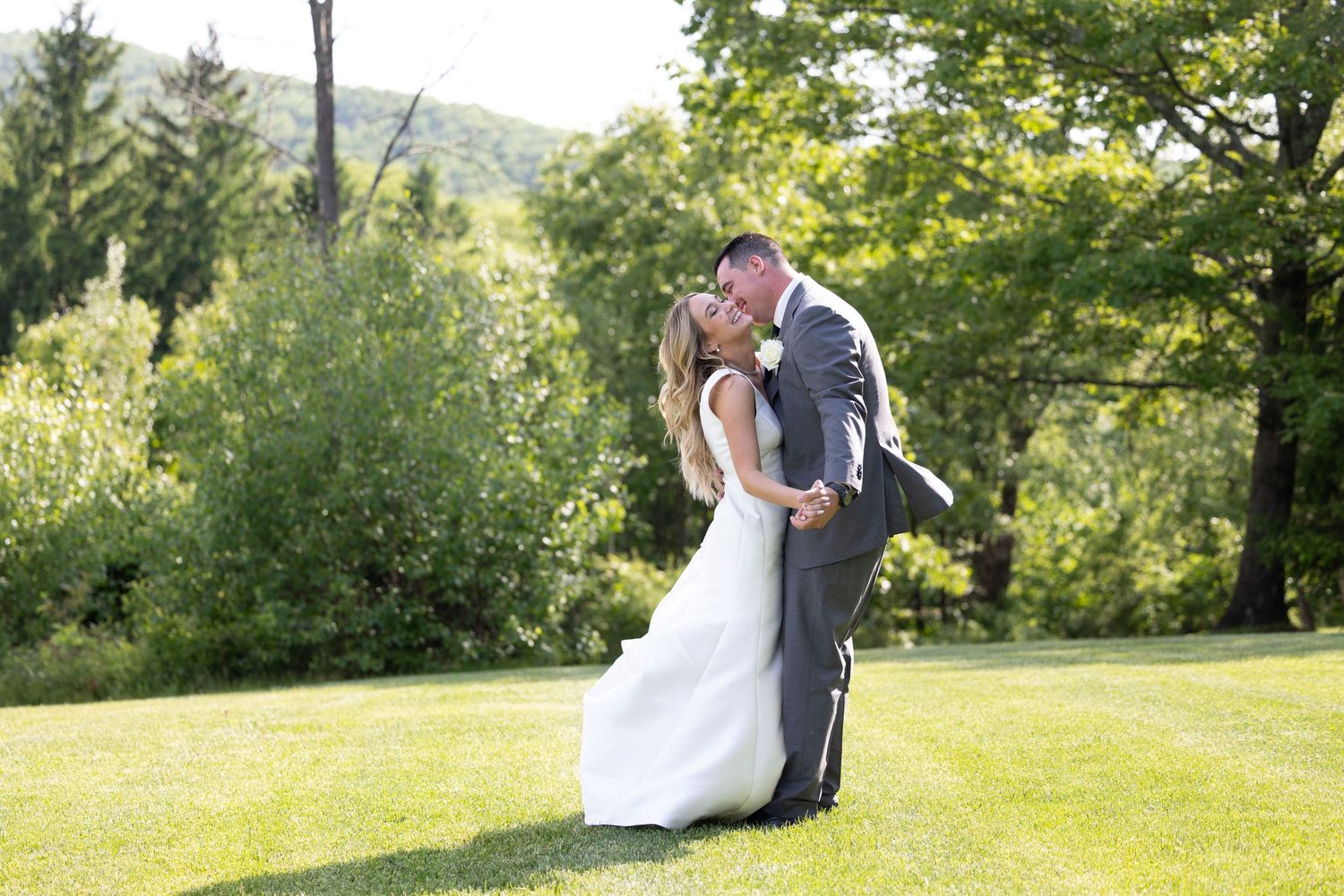 A couple in wedding attire share an intimate moment on a lush green lawn surrounded by trees on a sunny day.