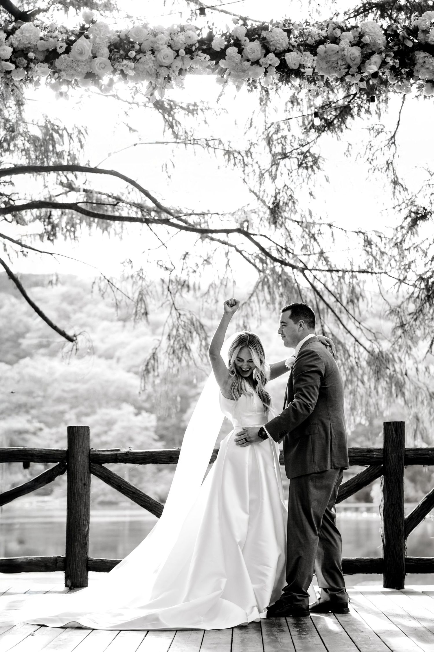 A romantic black and white wedding photo of a couple dancing on a wooden deck surrounded by overhanging trees.