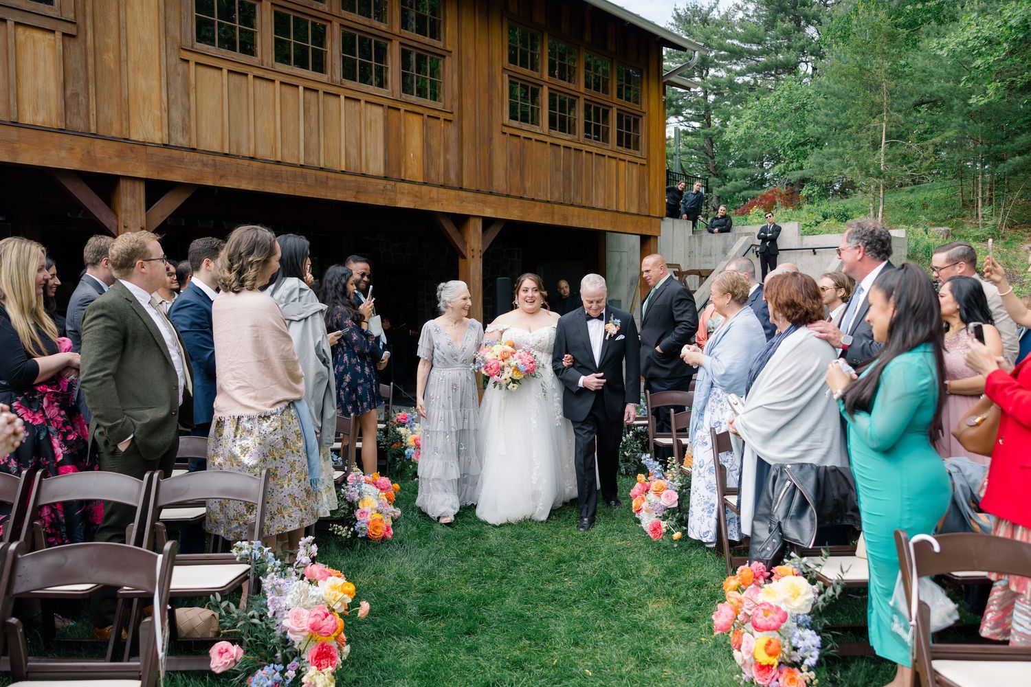 Wedding ceremony gathering outside rustic wooden building with guests watching celebration.