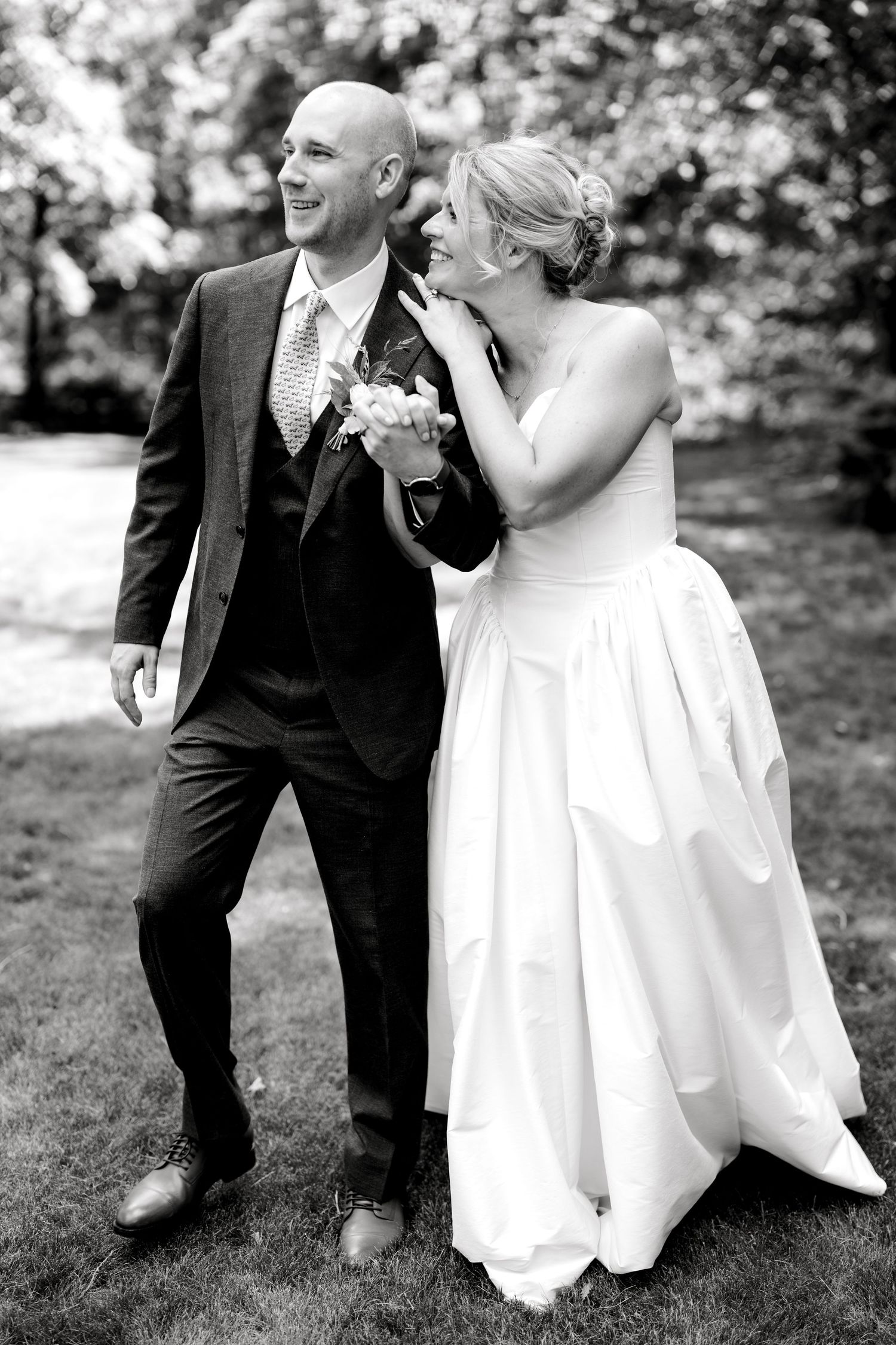 Black and white photo of a laughing bride in white gown whispering to her bald groom in a dark suit outdoors.