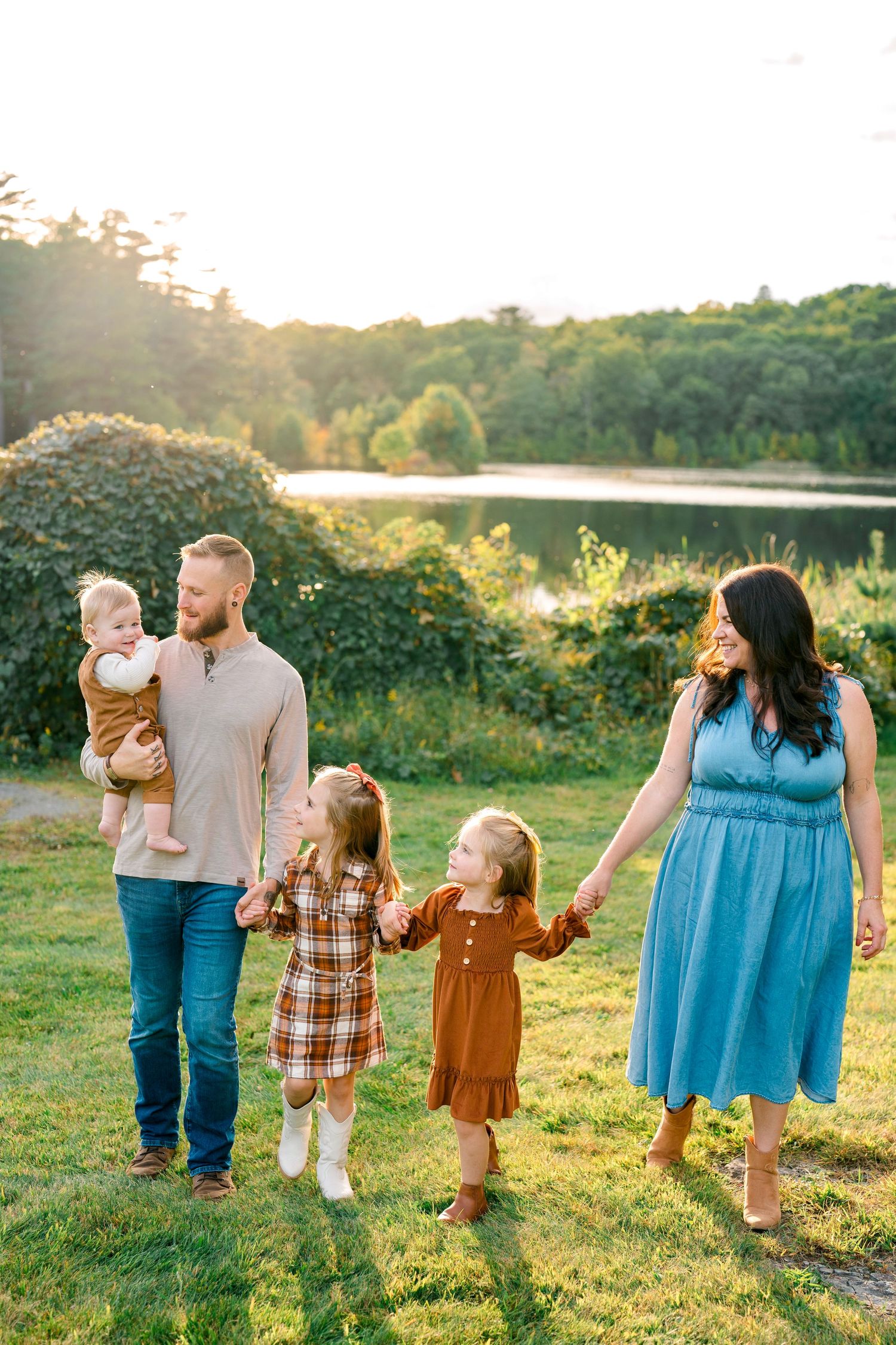 Family of five walking outdoors near a lake at golden hour, parents holding hands with young children in autumn outfits.