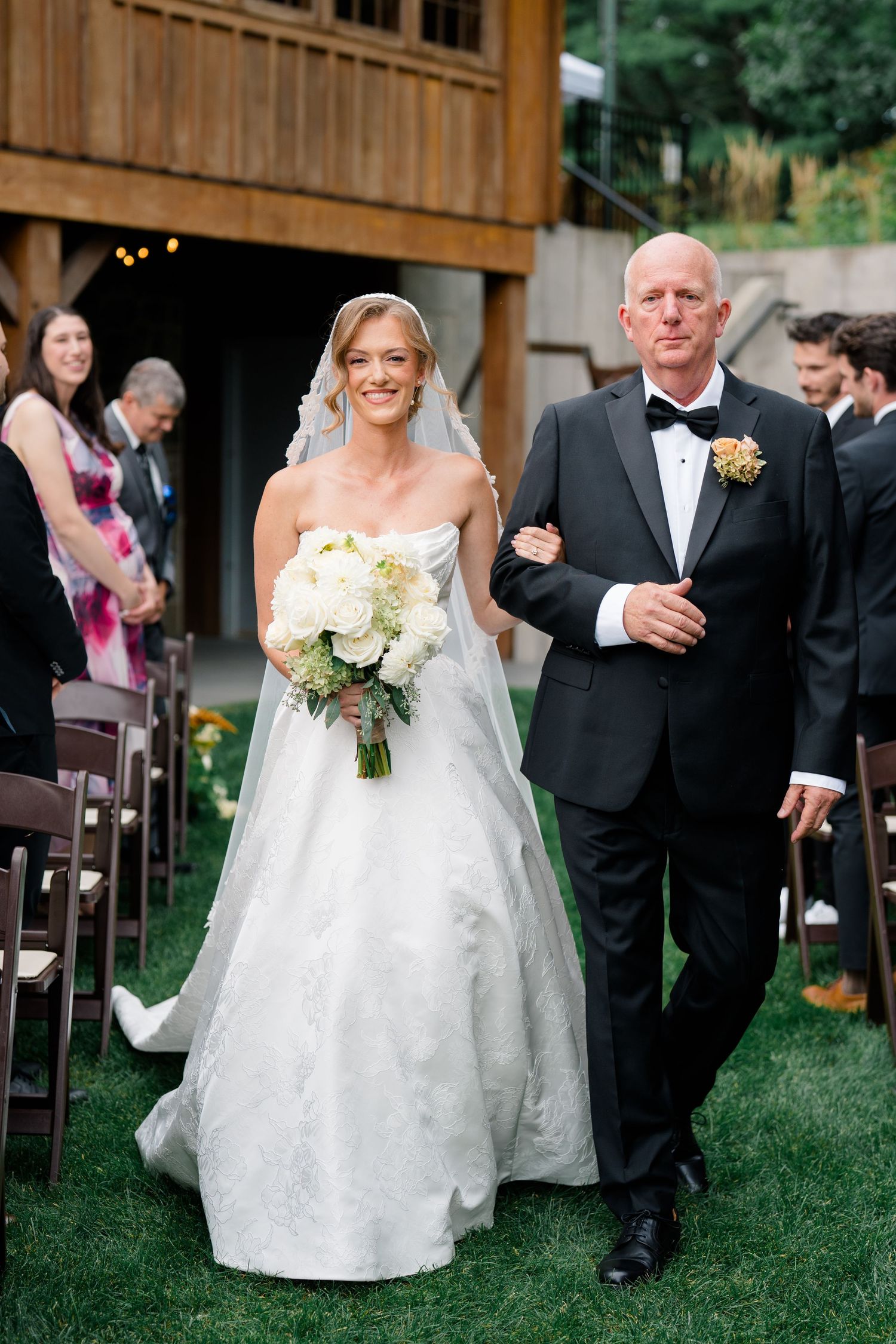Bride in white ball gown walks down aisle with father in tuxedo at outdoor wedding ceremony.