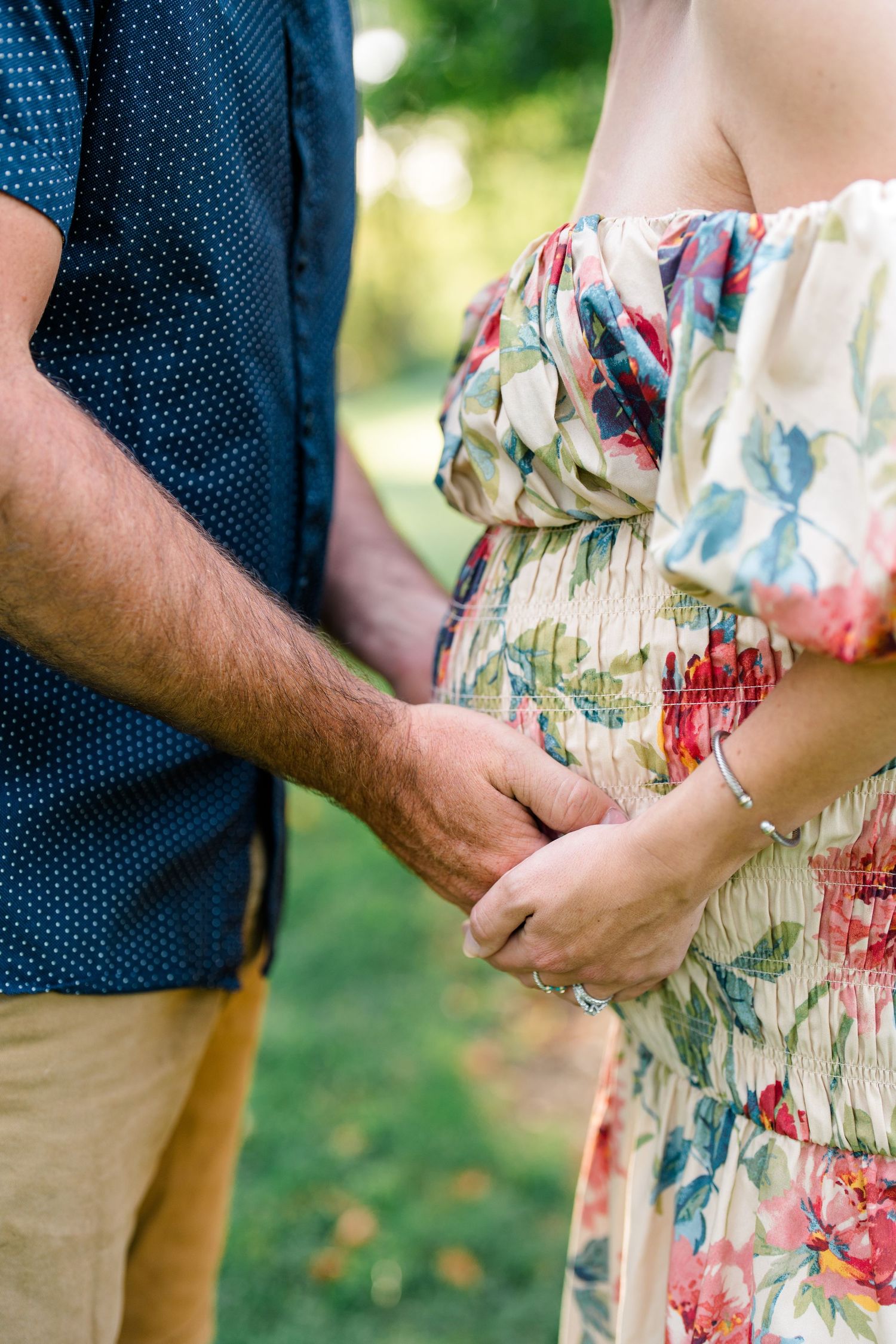 Couple holding hands outdoors, pregnant woman in floral off-shoulder dress with partner in blue shirt.