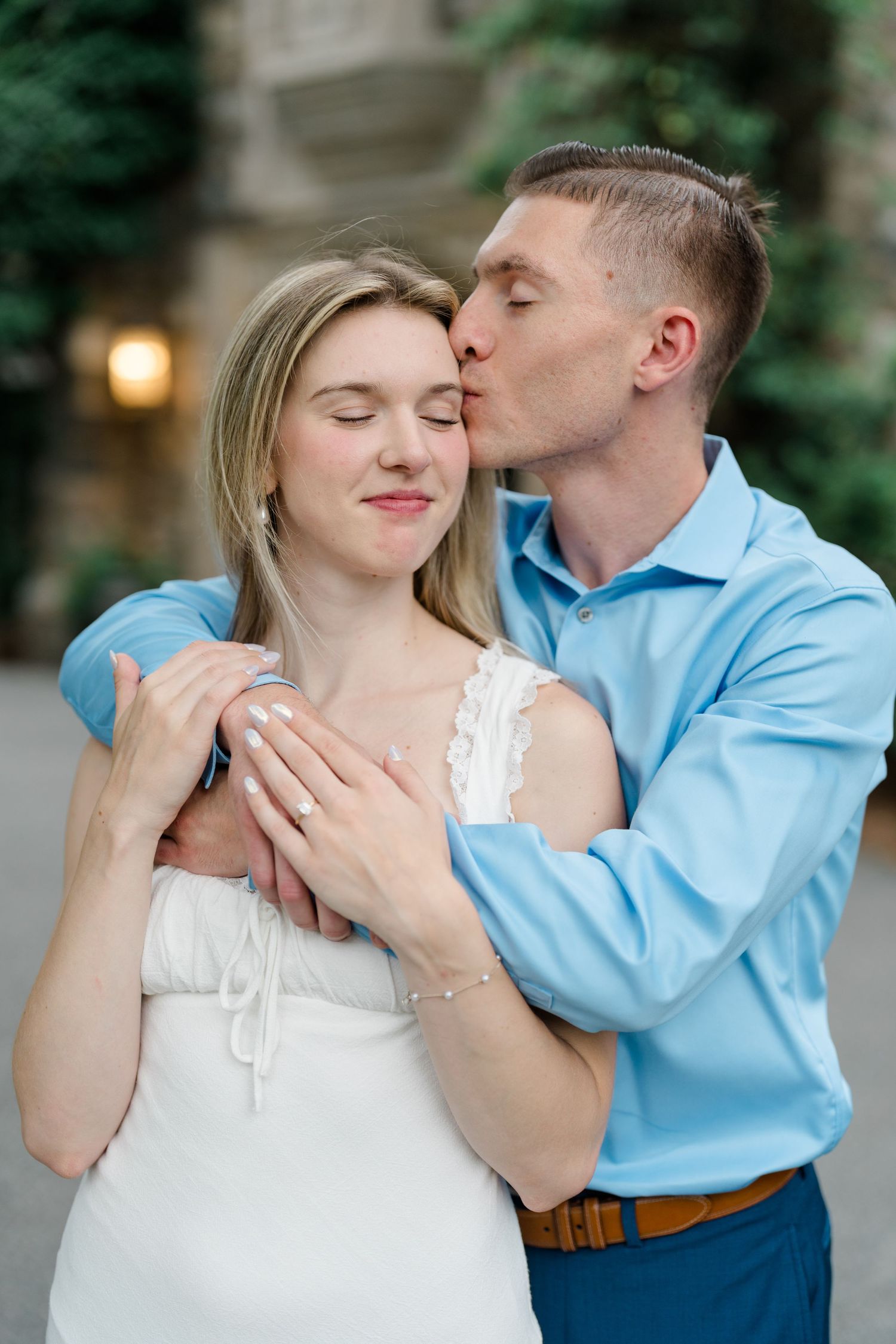 A man in a blue shirt tenderly kisses a woman in a white dress on the cheek while embracing her outdoors.