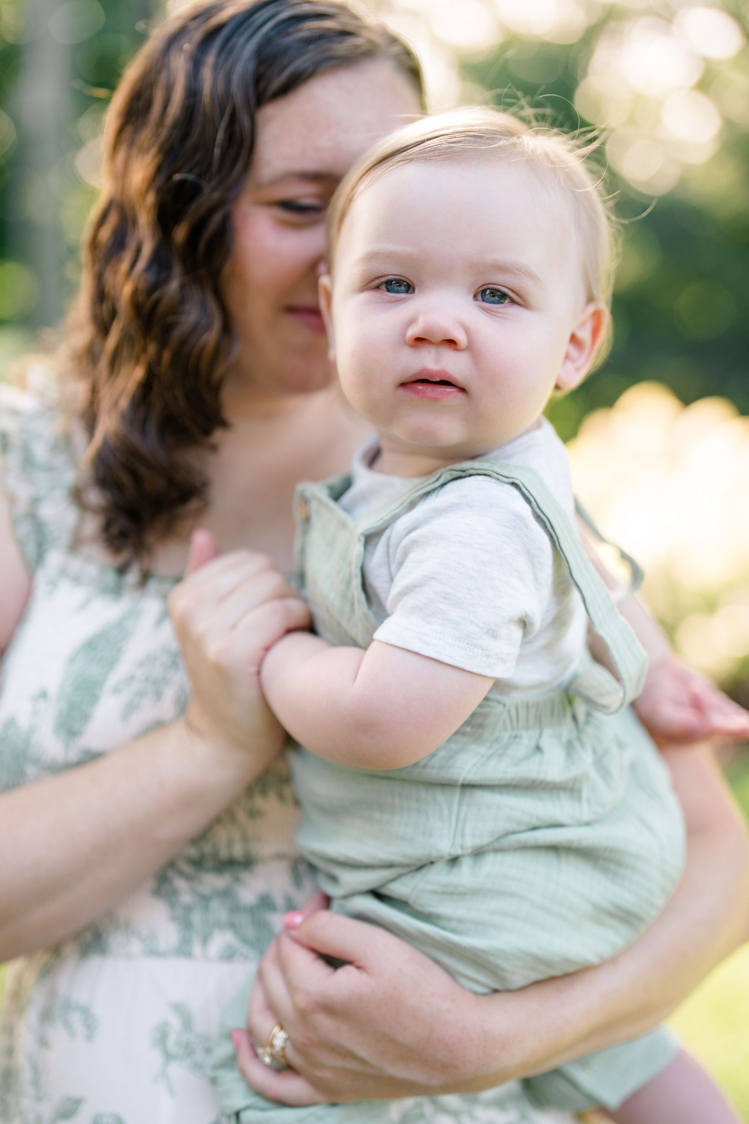 Mother holding her baby boy in a green romper outdoors in soft natural light.