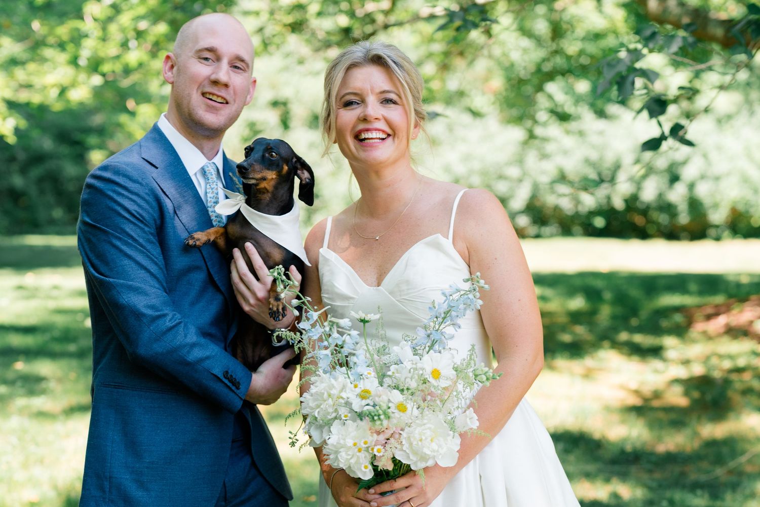 Smiling bride and groom in blue suit hold a dachshund puppy outdoors, bride carries white daisy bouquet.