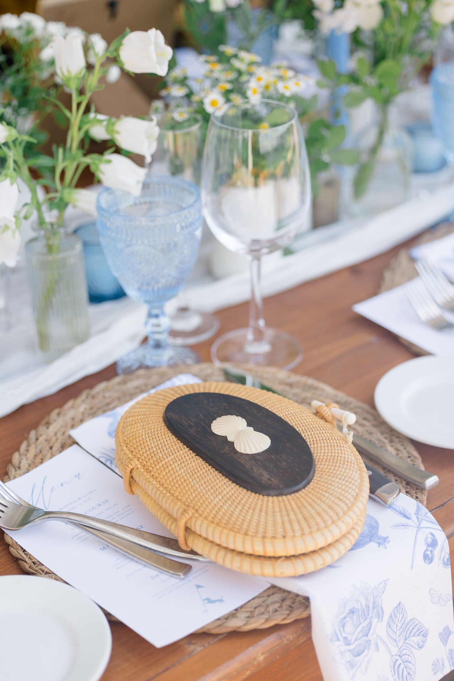Elegant wedding tablescape with wicker charger plates, blue glassware, and white daisy floral centerpieces.