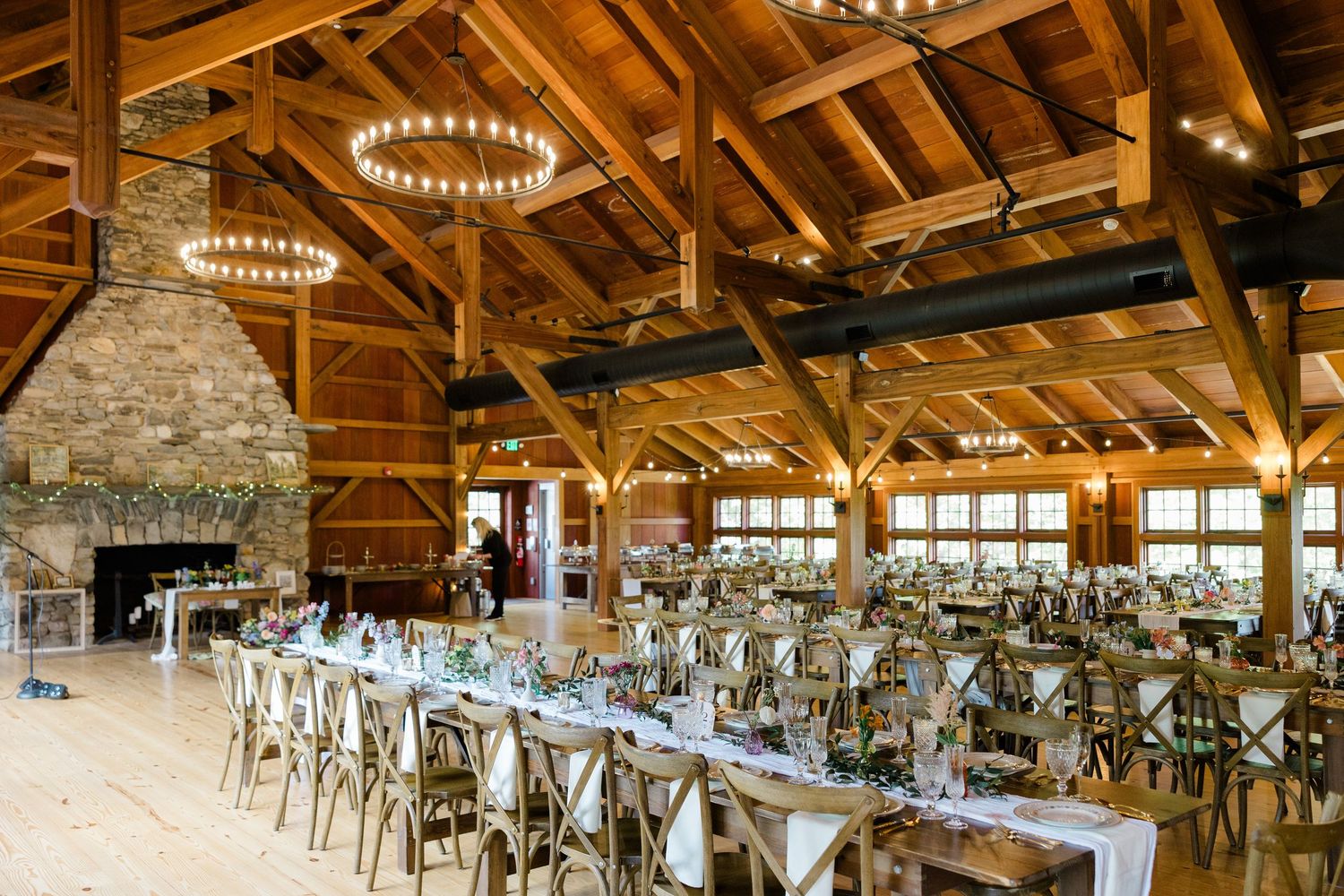 Rustic wedding reception venue with exposed wooden beams, chandeliers and long tables set for dining in a barn-style interior.