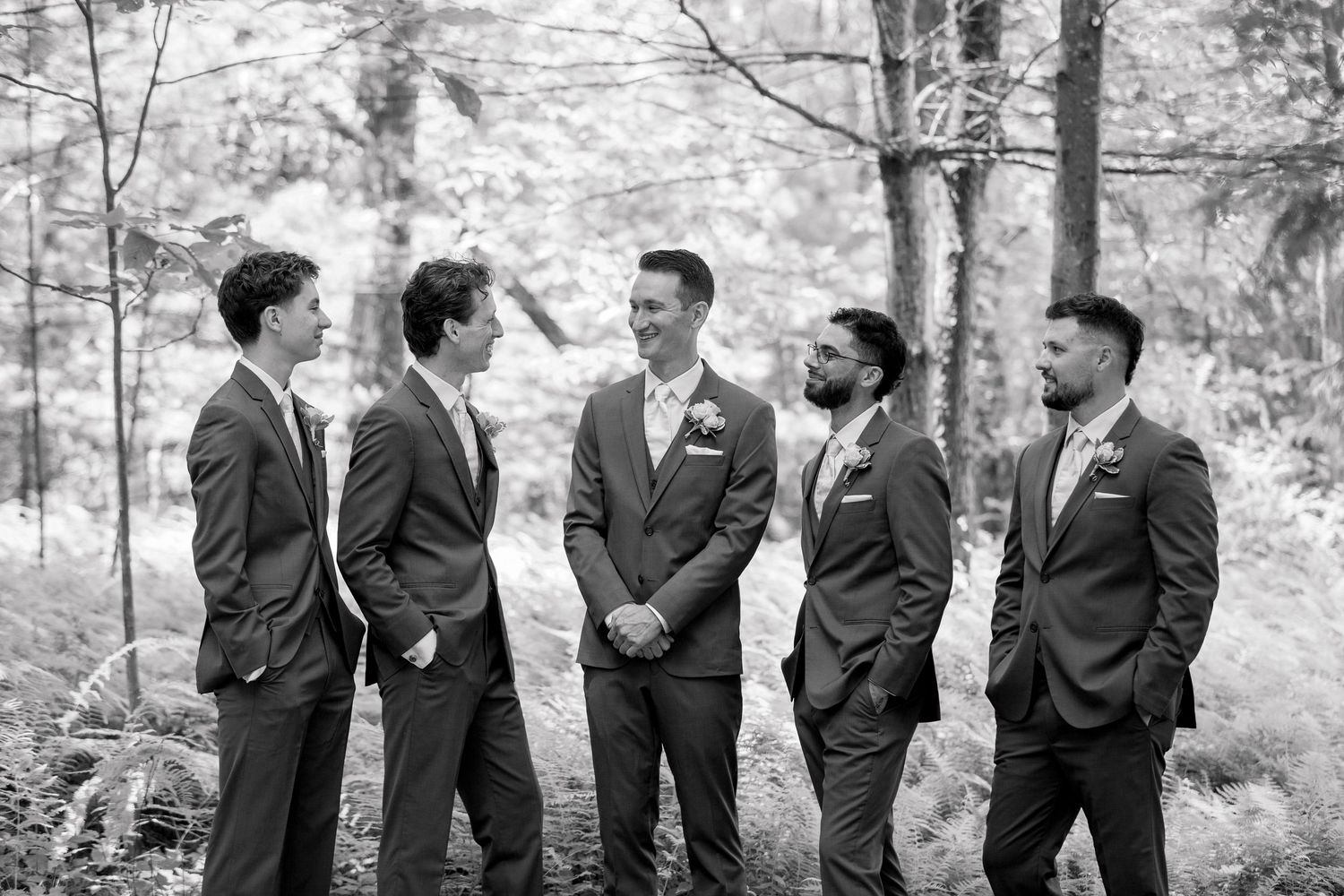 Groomsmen in dark suits line up for black and white photo in forest setting.