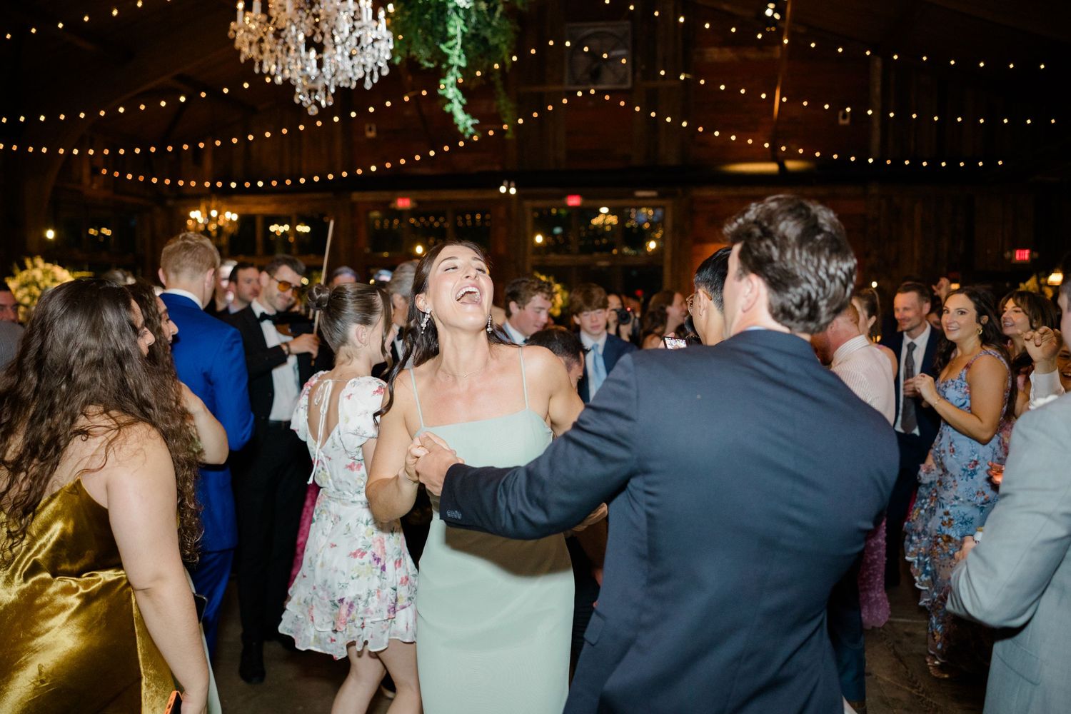 Wedding guests dancing and celebrating under twinkling lights and chandeliers at an indoor reception.