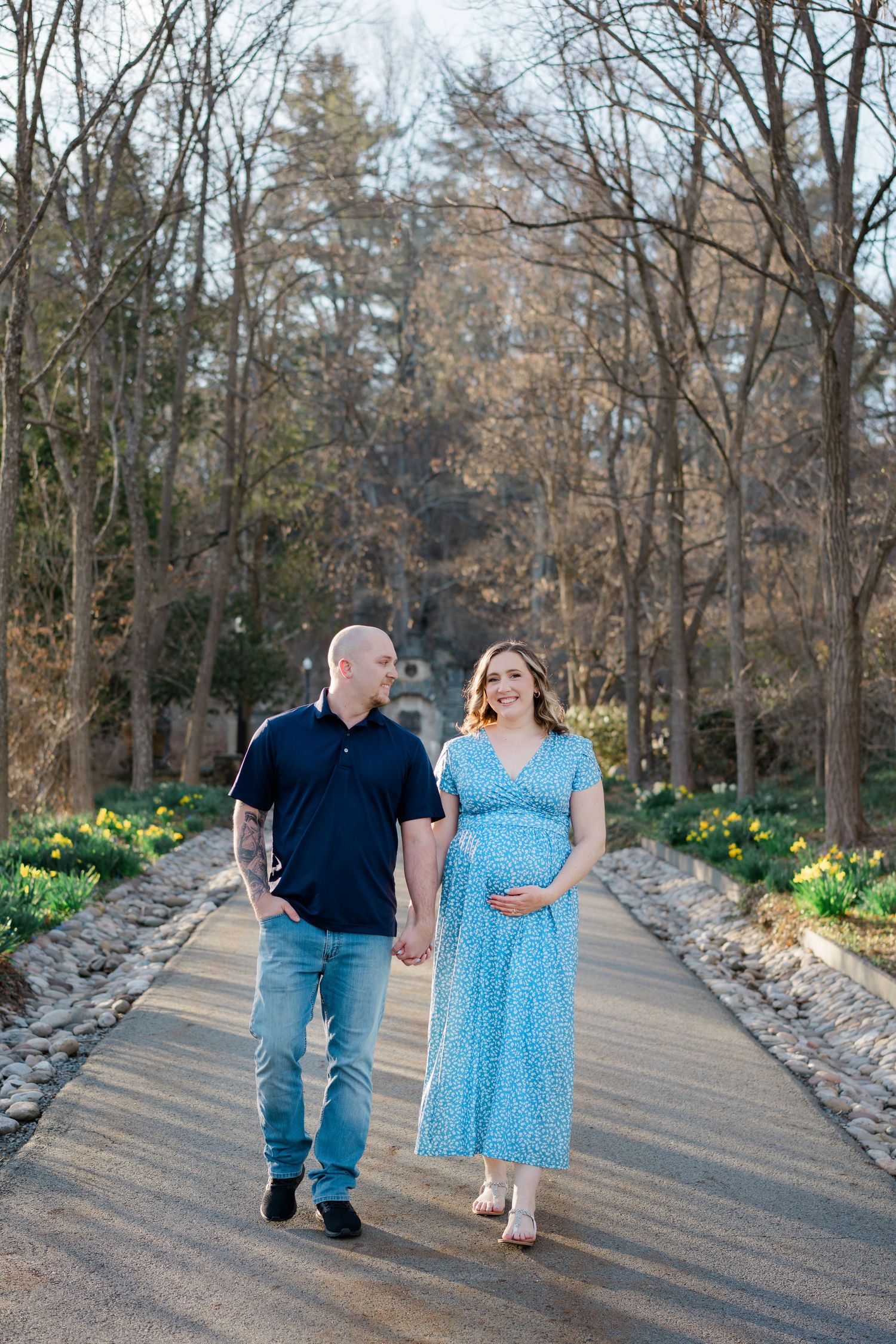 A couple walks hand in hand down a tree-lined path with daffodils blooming along the edges during springtime.