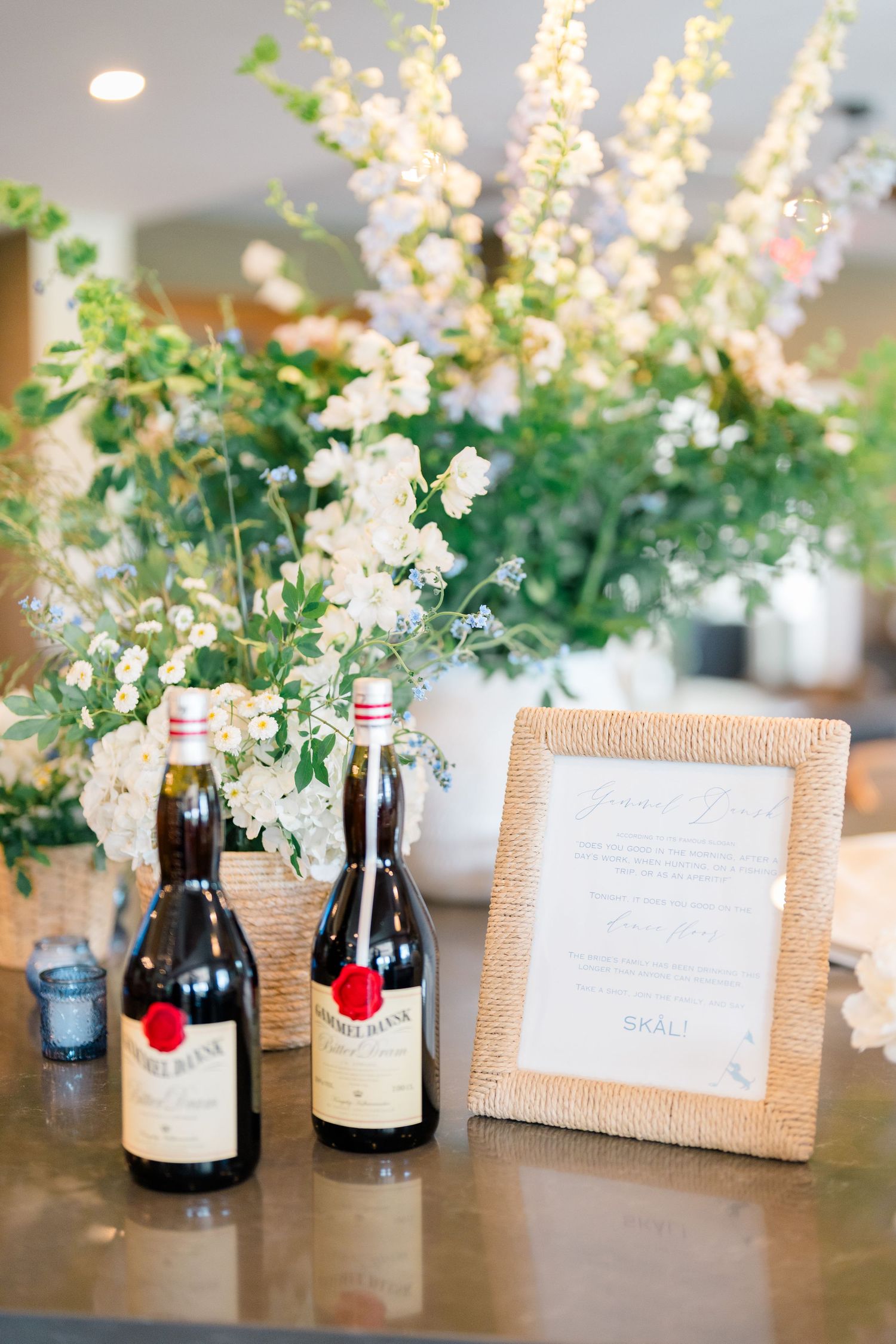 Wedding reception bar display with wine bottles, framed menu, and large white floral arrangement.