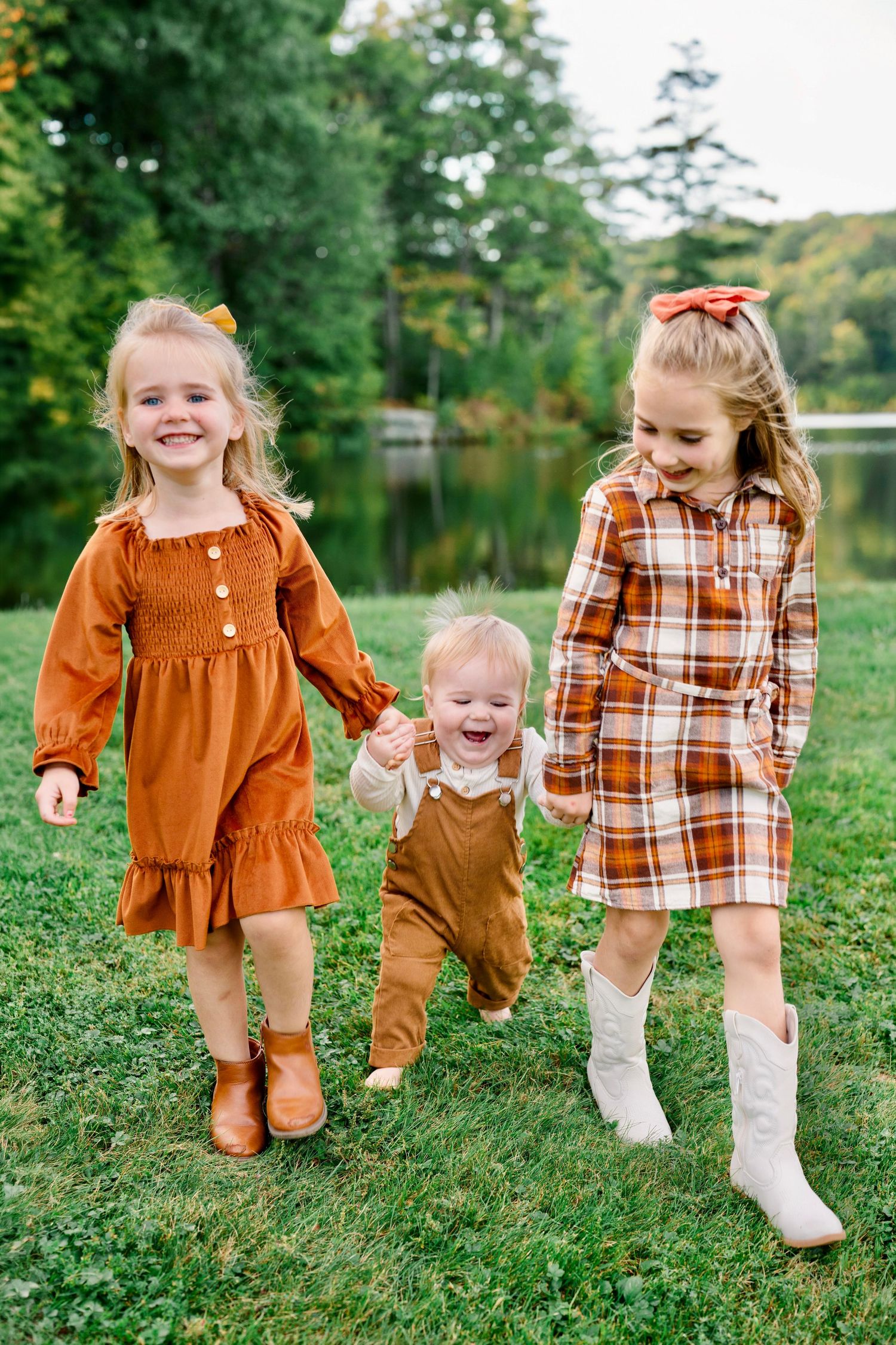 Three young siblings in autumn outfits walk together on grass near a lake, with two girls holding a laughing baby boy.