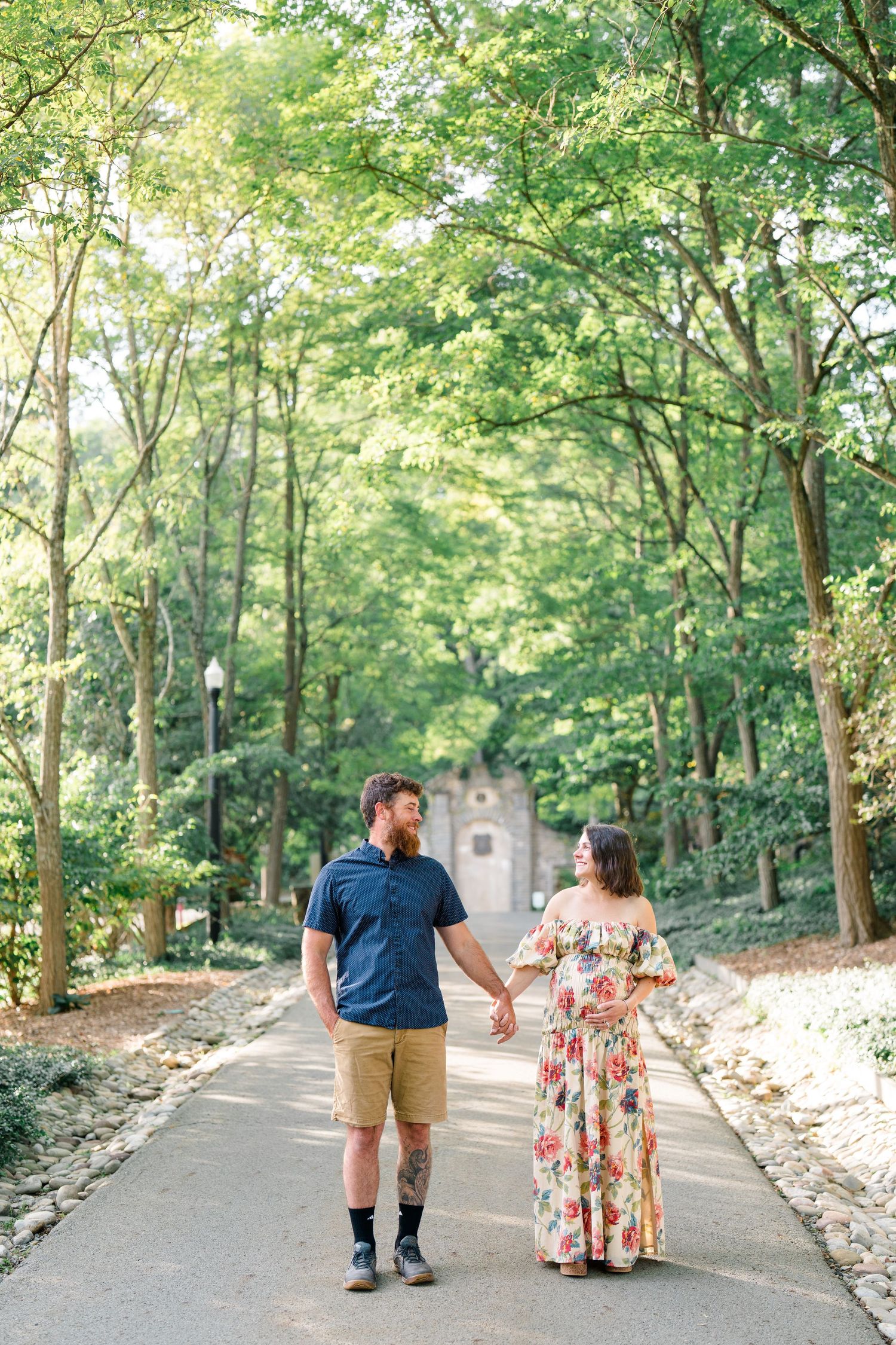 Couple holding hands walking down a tree-lined path, man in navy shirt and khaki shorts, woman in floral maxi dress.