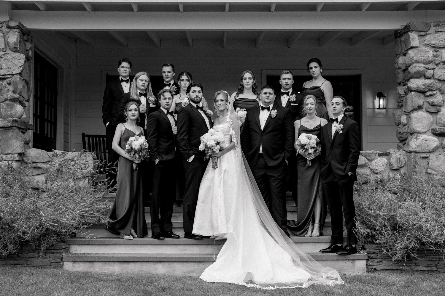 Black and white wedding party photo on stone steps, bride in gown with veil, groom and groomsmen in tuxedos, bridesmaids in gowns.