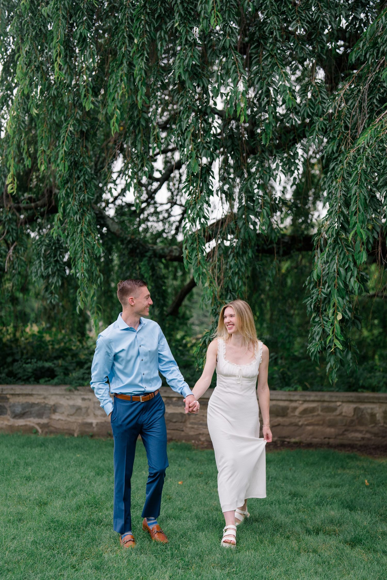 Smiling couple holding hands and walking on green grass under a weeping willow tree, man in blue shirt and woman in white dress.
