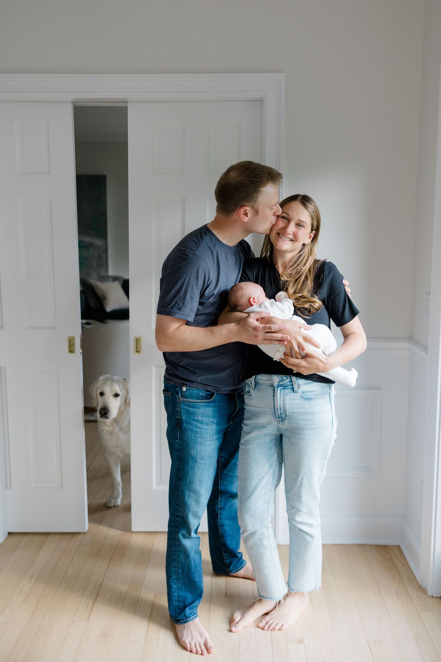 Family shares tender moment in hallway while golden retriever peeks from doorway.