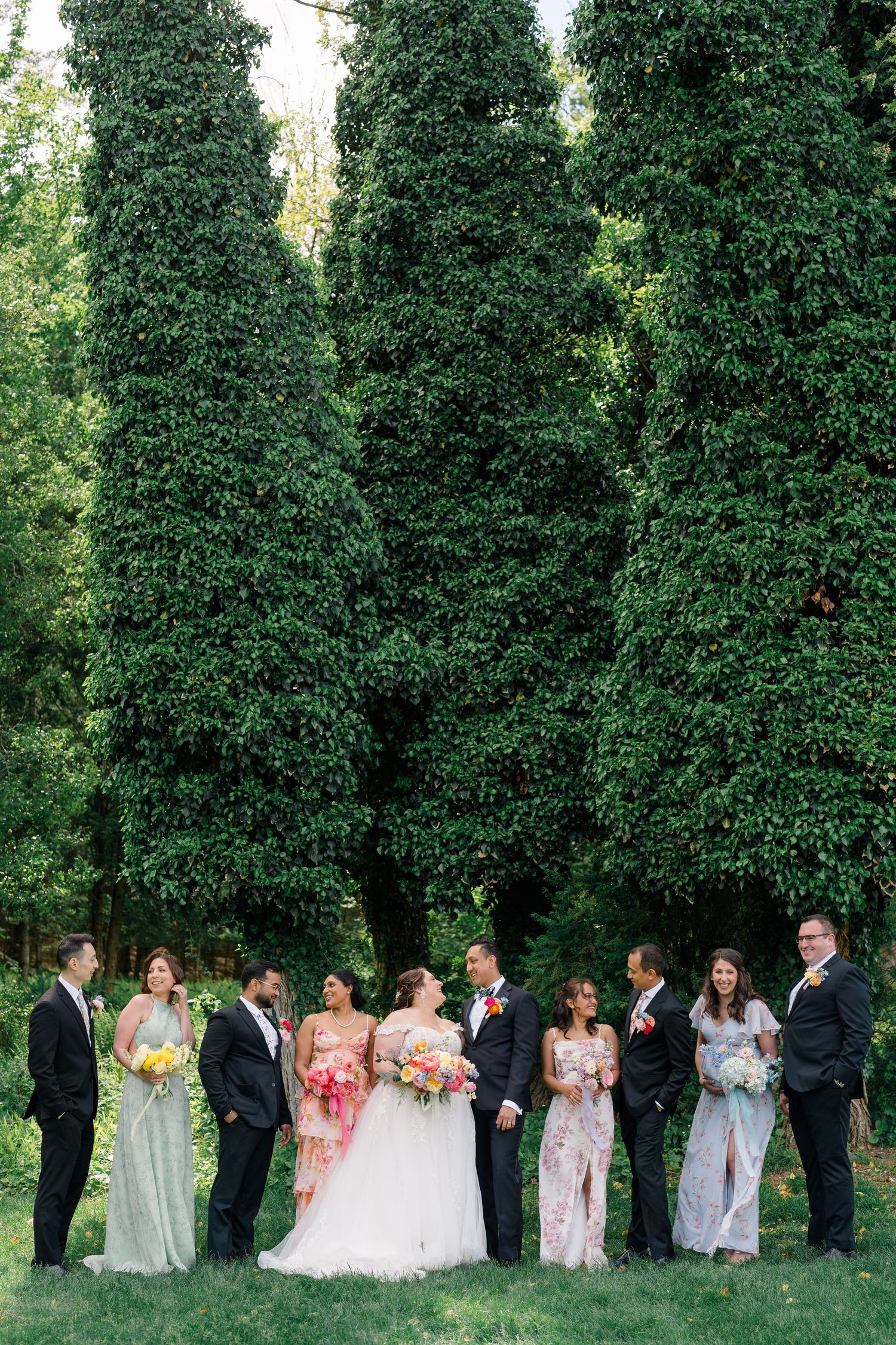 Wedding party poses against tall green trees in an outdoor garden setting.