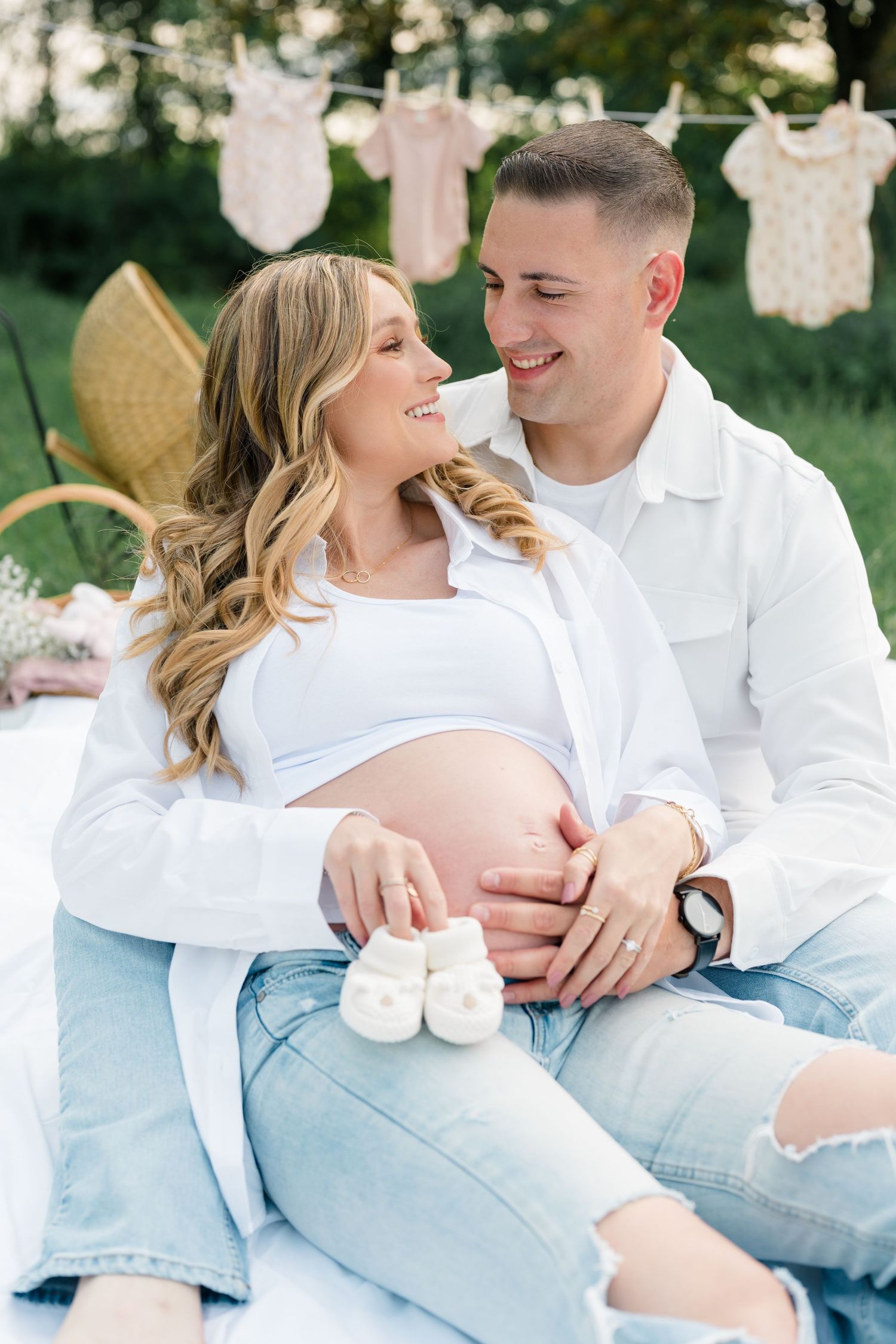 A maternity photoshoot outdoors with baby clothes on clothesline and wicker chair as props.