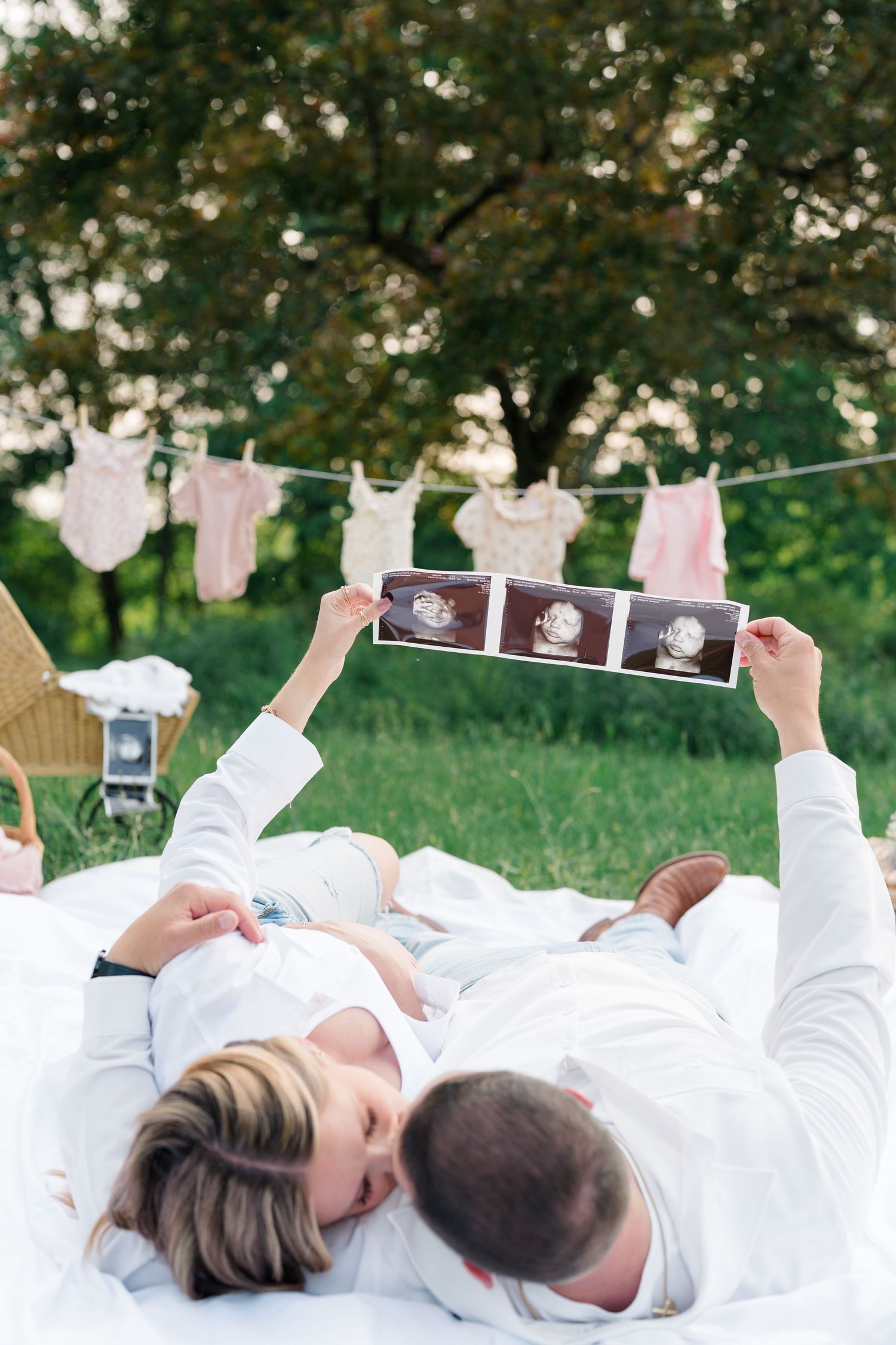 Couple laying on white blanket outdoors holding ultrasound photos in a creative pregnancy announcement.