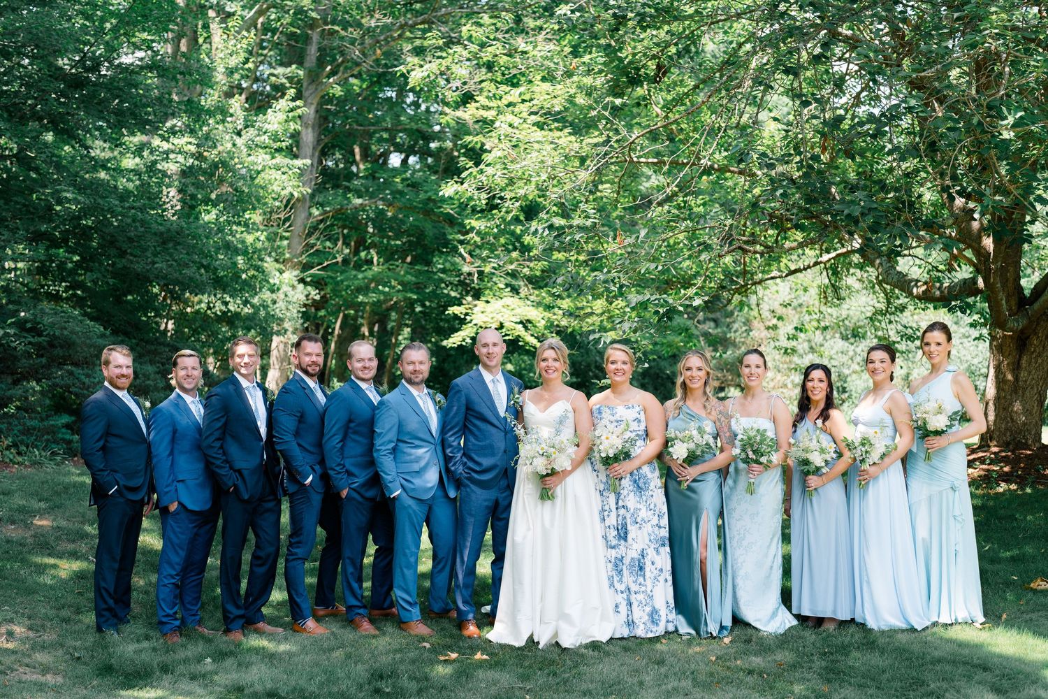 Wedding party in blue suits and light blue bridesmaid dresses posing outdoors among lush green trees.