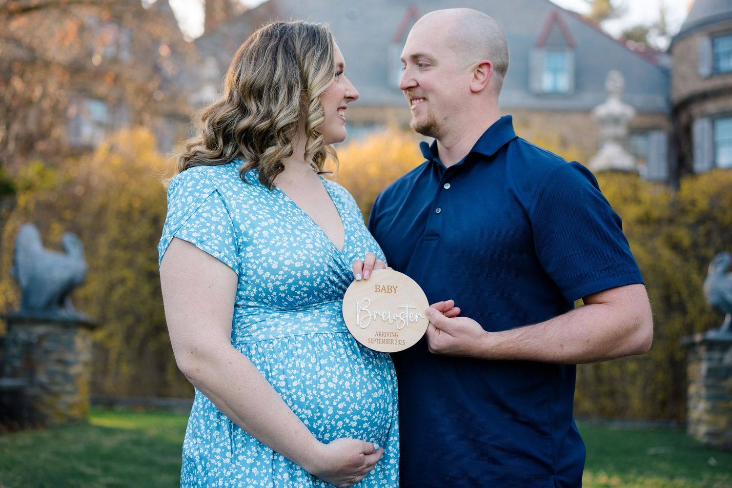 A couple celebrates their pregnancy in an outdoor setting, with the woman wearing a blue dress and holding a wooden date marker.