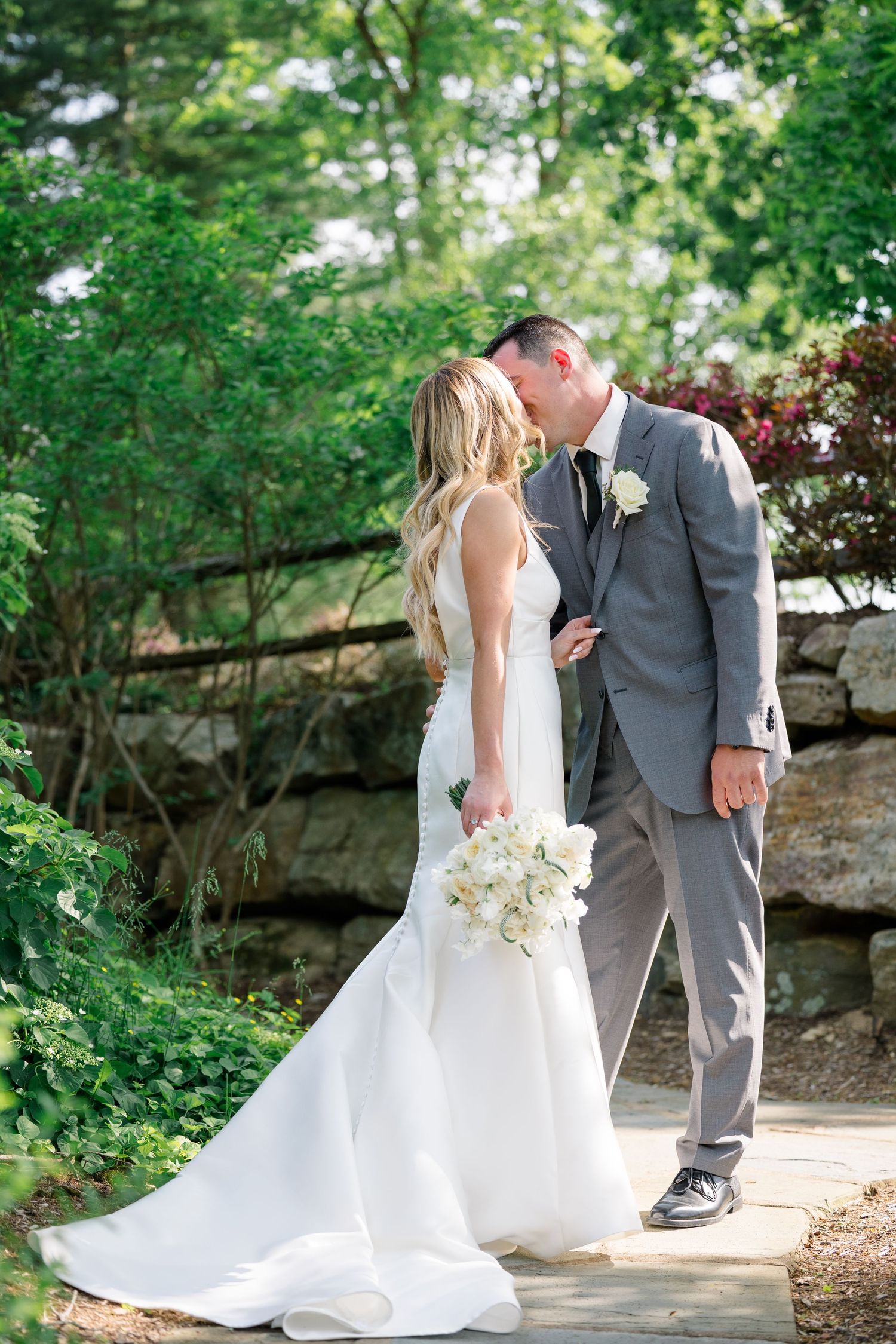 A romantic wedding couple shares a tender moment on a stone path surrounded by lush green gardens.