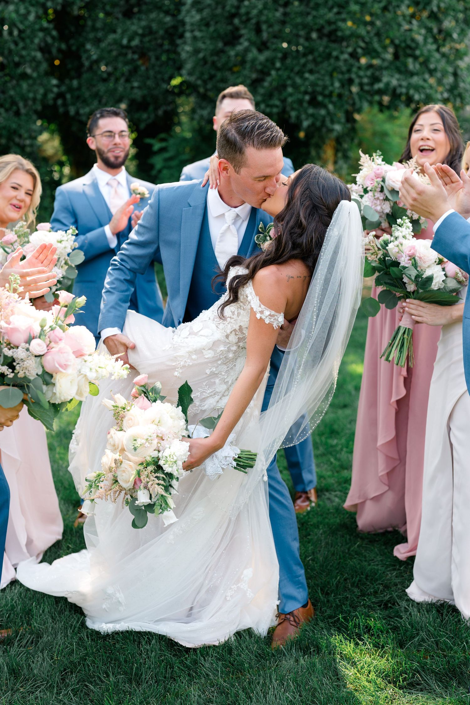 Wedding party in blue suits and pink dresses laughing together on grassy lawn.