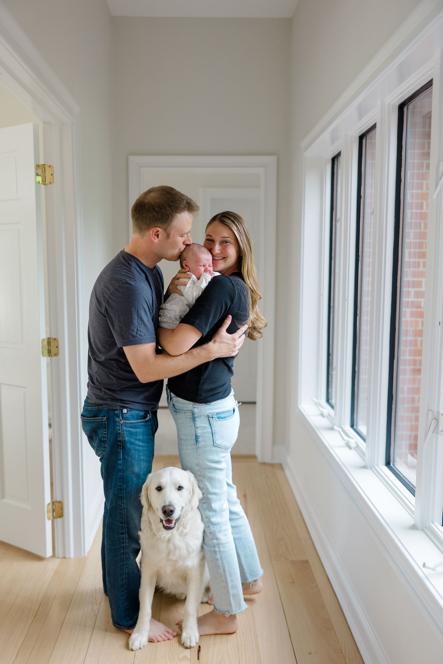 Family stands in hallway with golden retriever beside large windows.