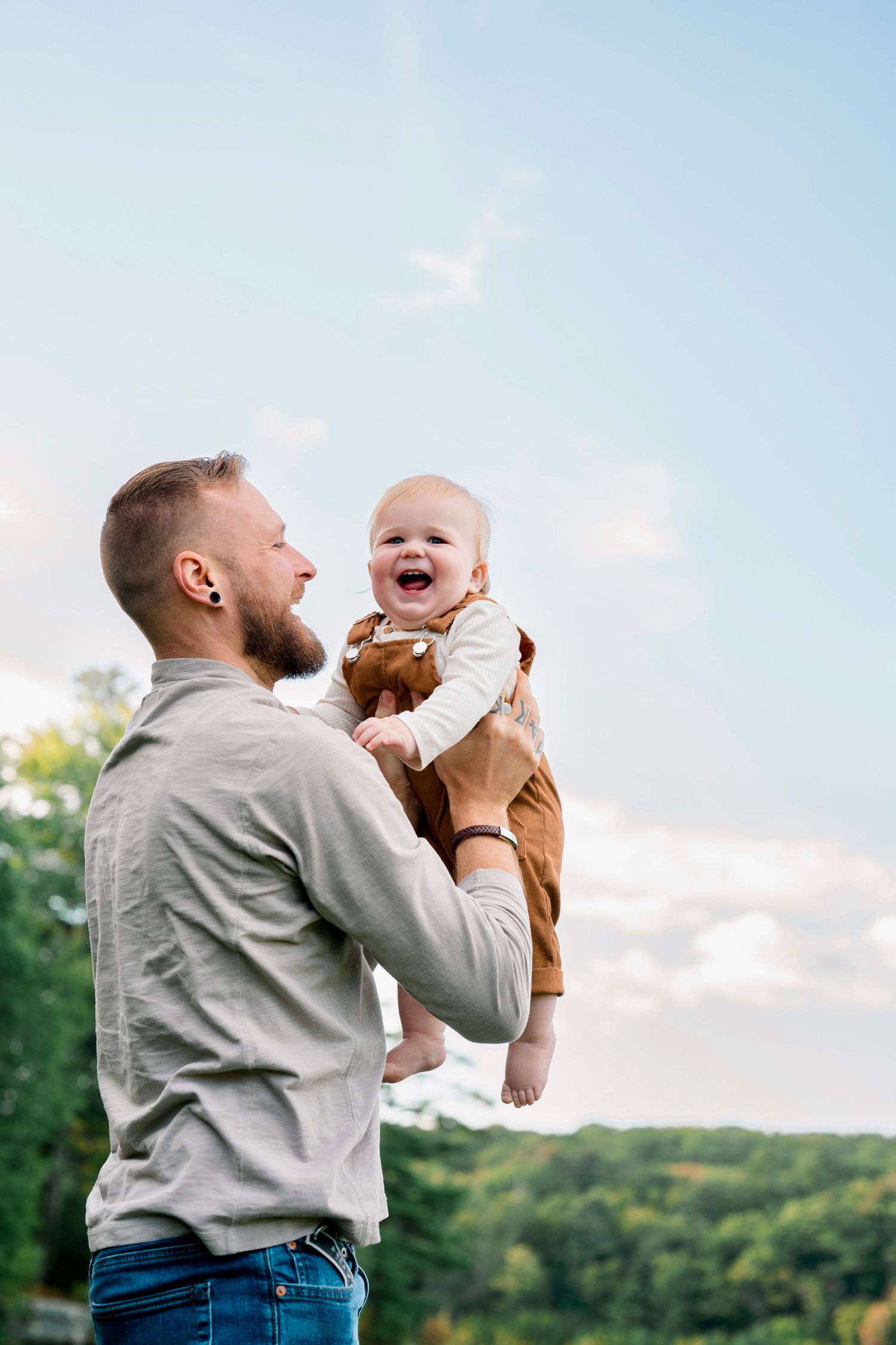 A bearded father joyfully lifts his laughing baby outdoors against a blue sky with green trees below.