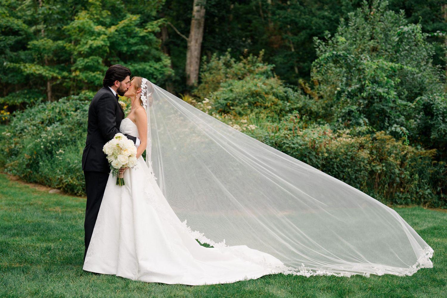 Bride and groom kissing outdoors with flowing cathedral veil billowing in the wind against lush green trees.
