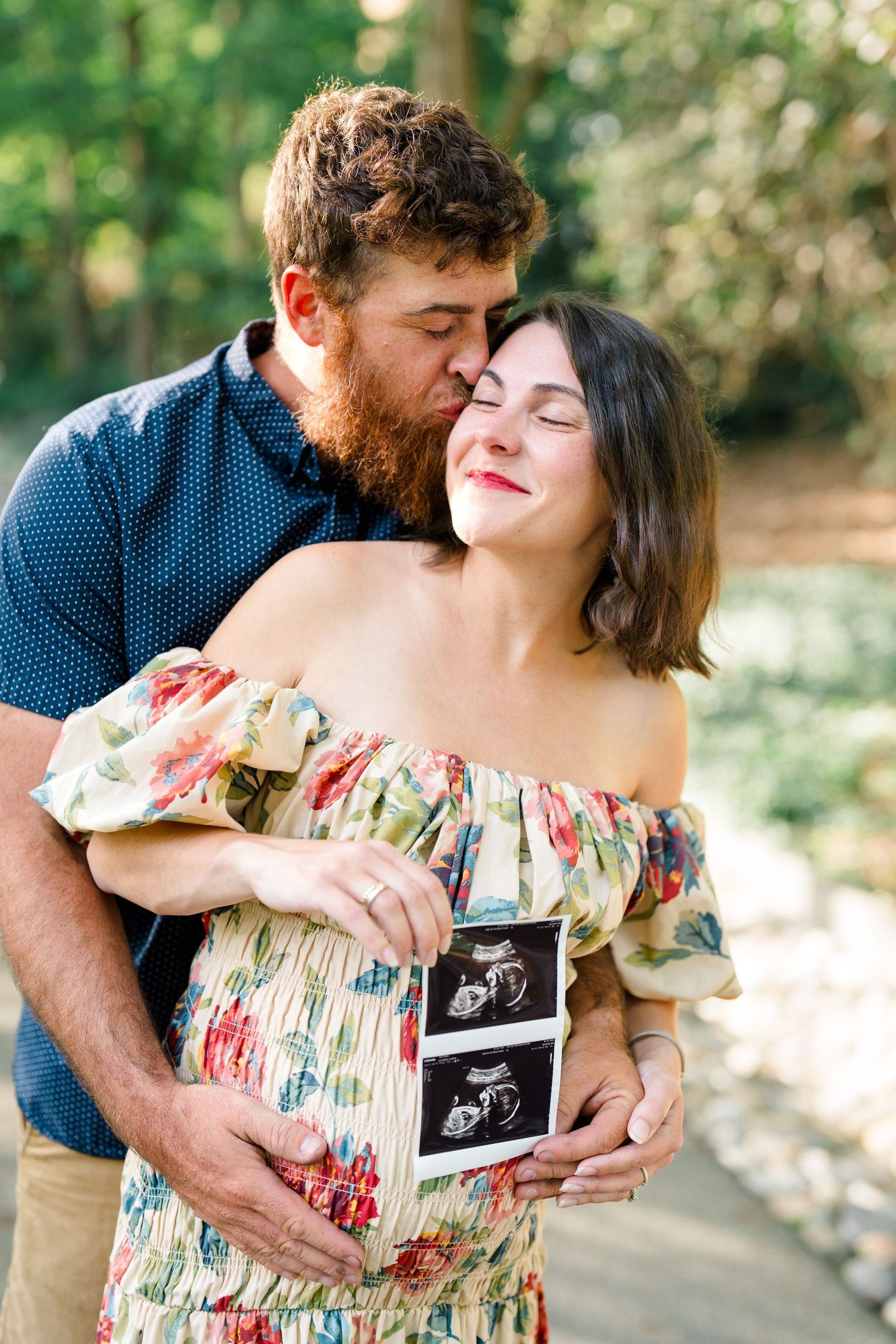 Pregnant woman in floral dress holds ultrasound photo as bearded man kisses her cheek outdoors.