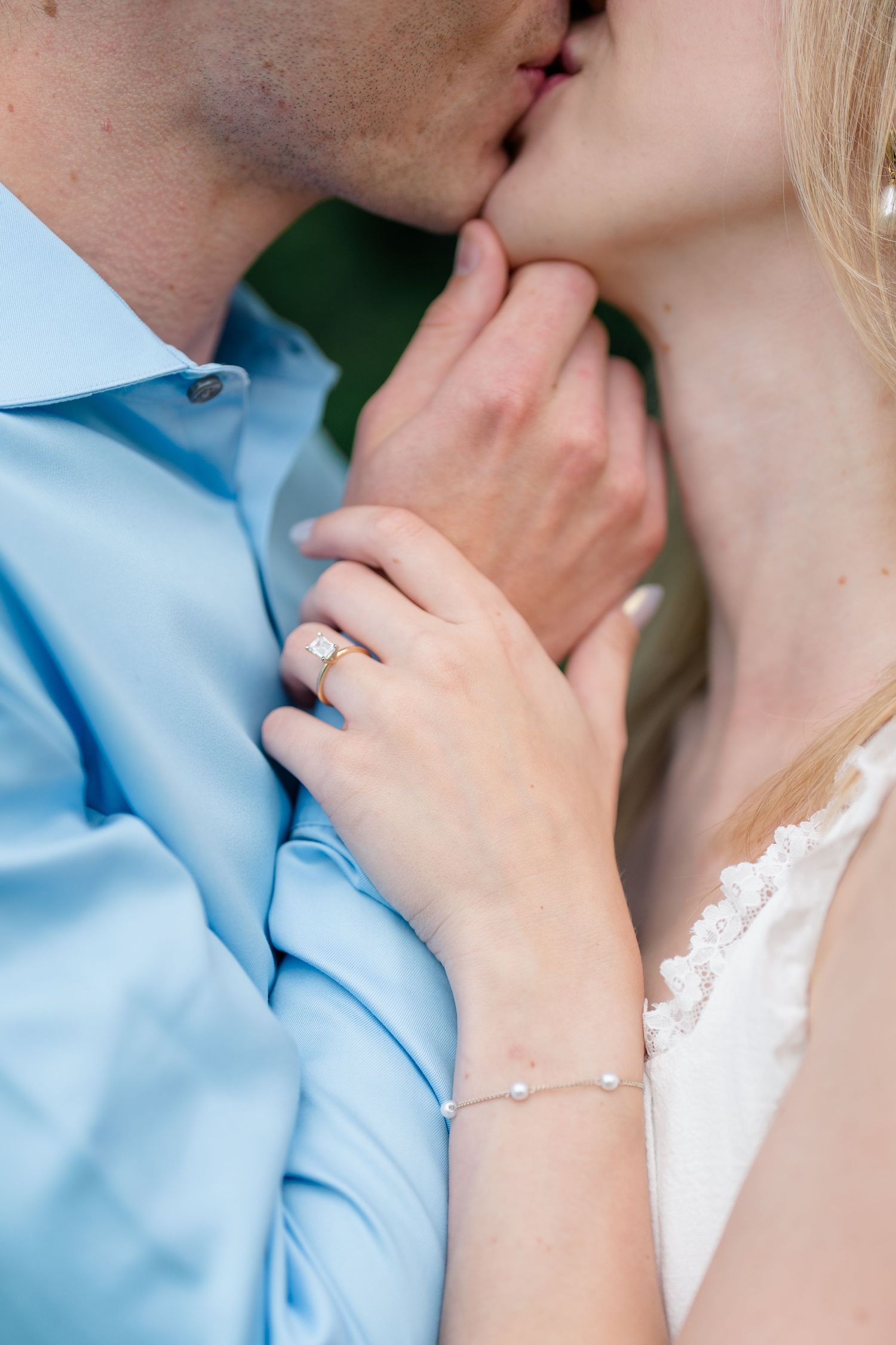 Close-up of couple kissing, woman displaying diamond engagement ring and pearl bracelet, man in light blue shirt.