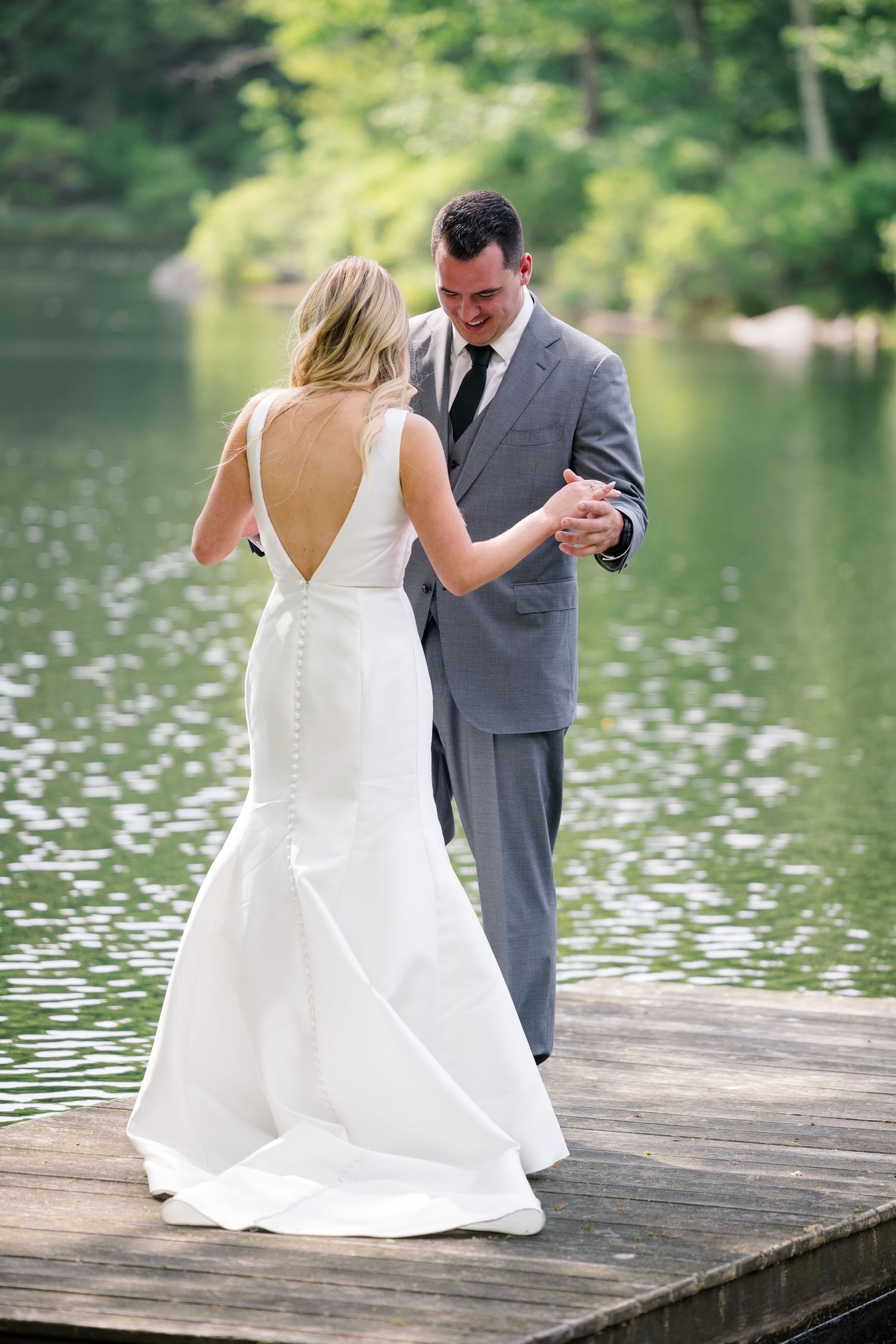 A bride in a white backless dress and groom in gray suit dancing together on a dock by a serene lake.