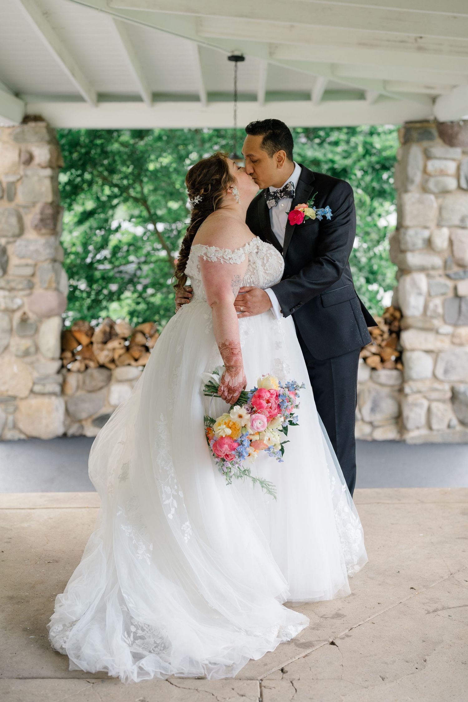 A romantic wedding couple sharing a tender moment under a stone pavilion with a vibrant floral bouquet.