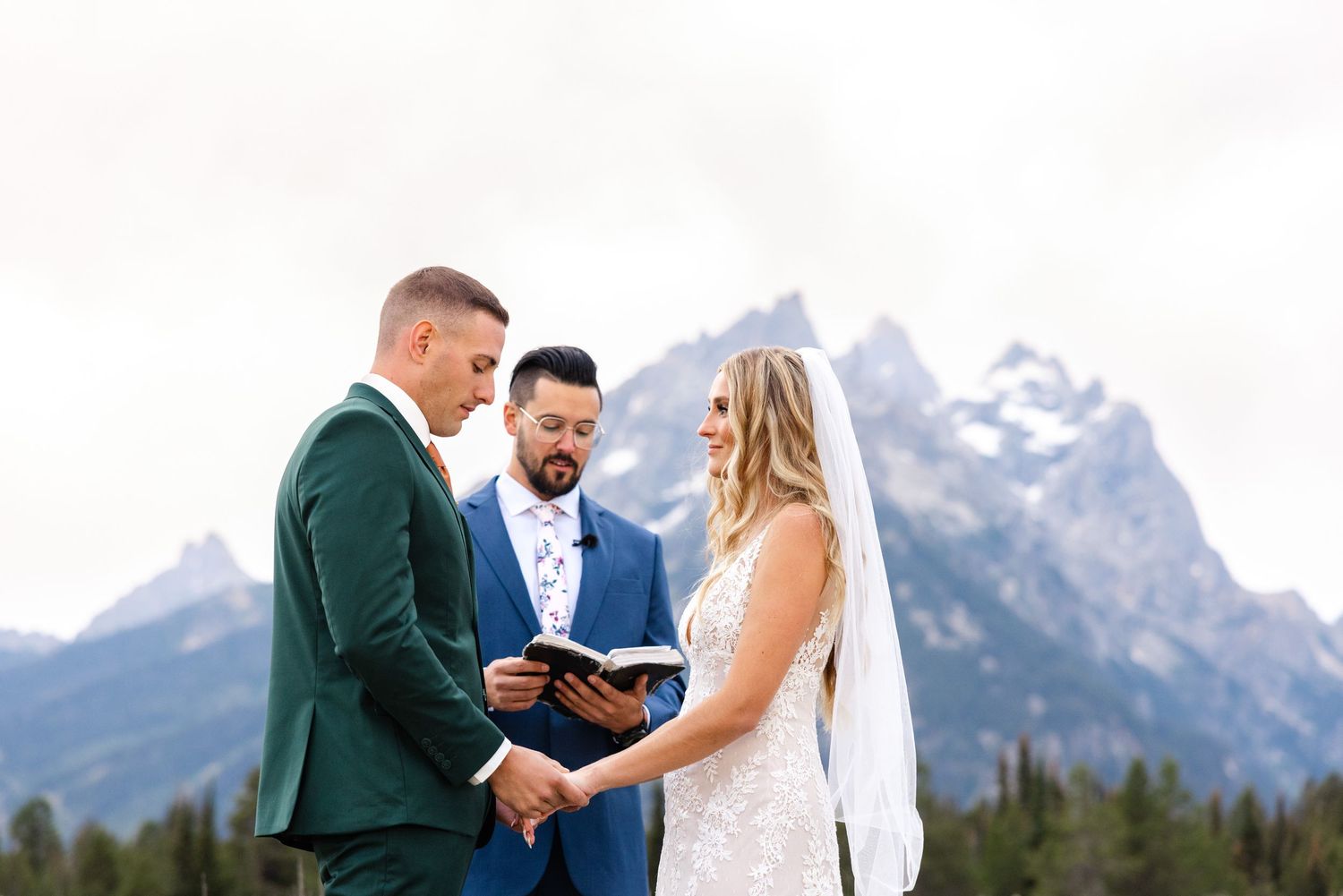 A couple exchanges vows during their outdoor wedding ceremony with snow-capped mountains and evergreen trees in the background.