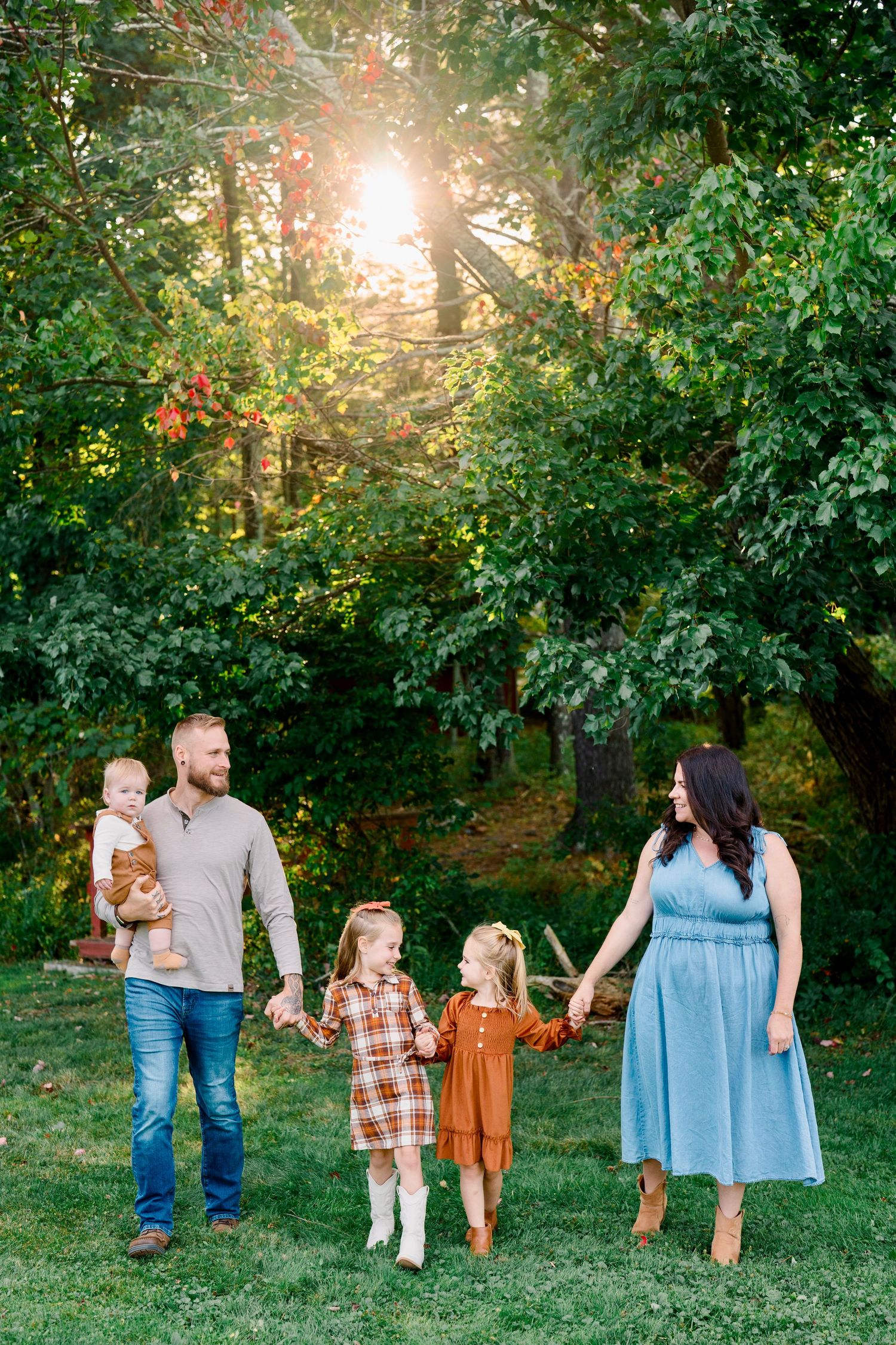 Family of five walking outdoors in golden sunlight, parents holding hands with two girls in fall outfits, dad carrying baby.