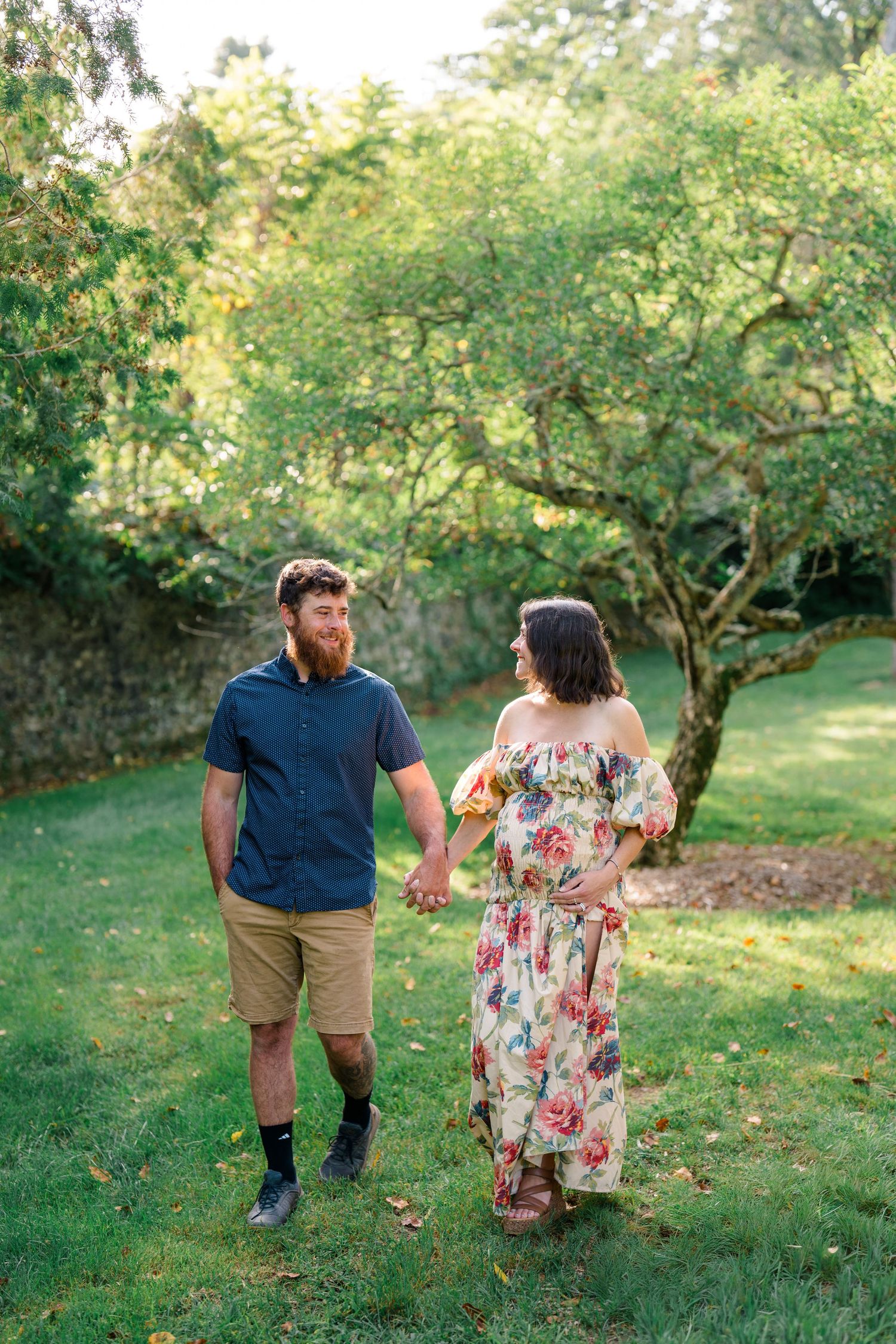 Couple holding hands walking through lush green garden, man with red beard in blue shirt and woman in floral maxi dress.