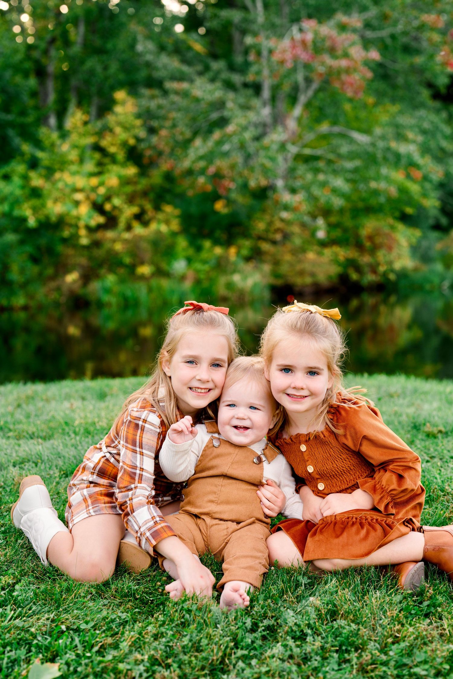 Three young sisters in autumn outfits sitting together on green grass, smiling for a fall family portrait outdoors.