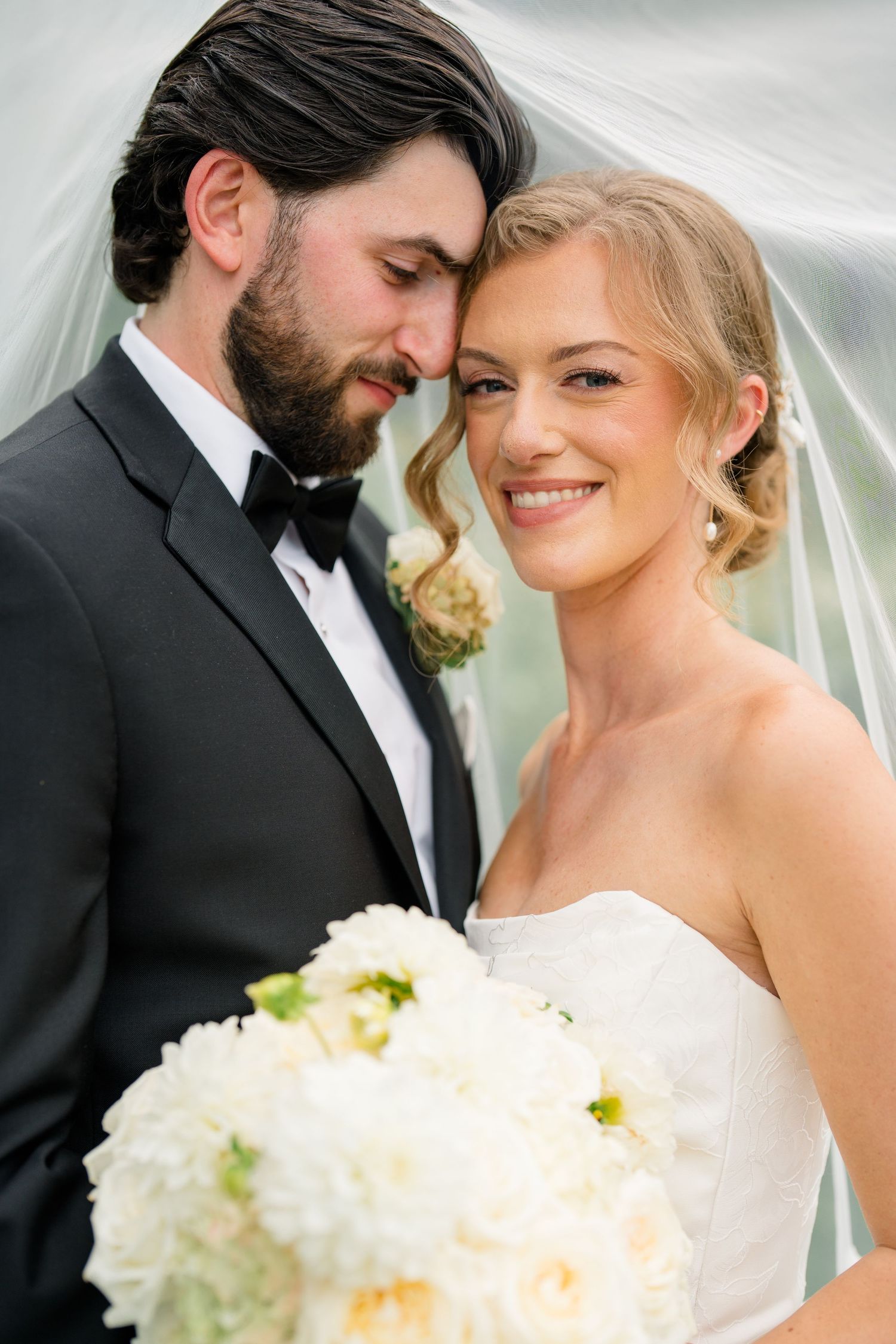 Close portrait of smiling bride with white bouquet leaning against bearded groom under flowing wedding veil.