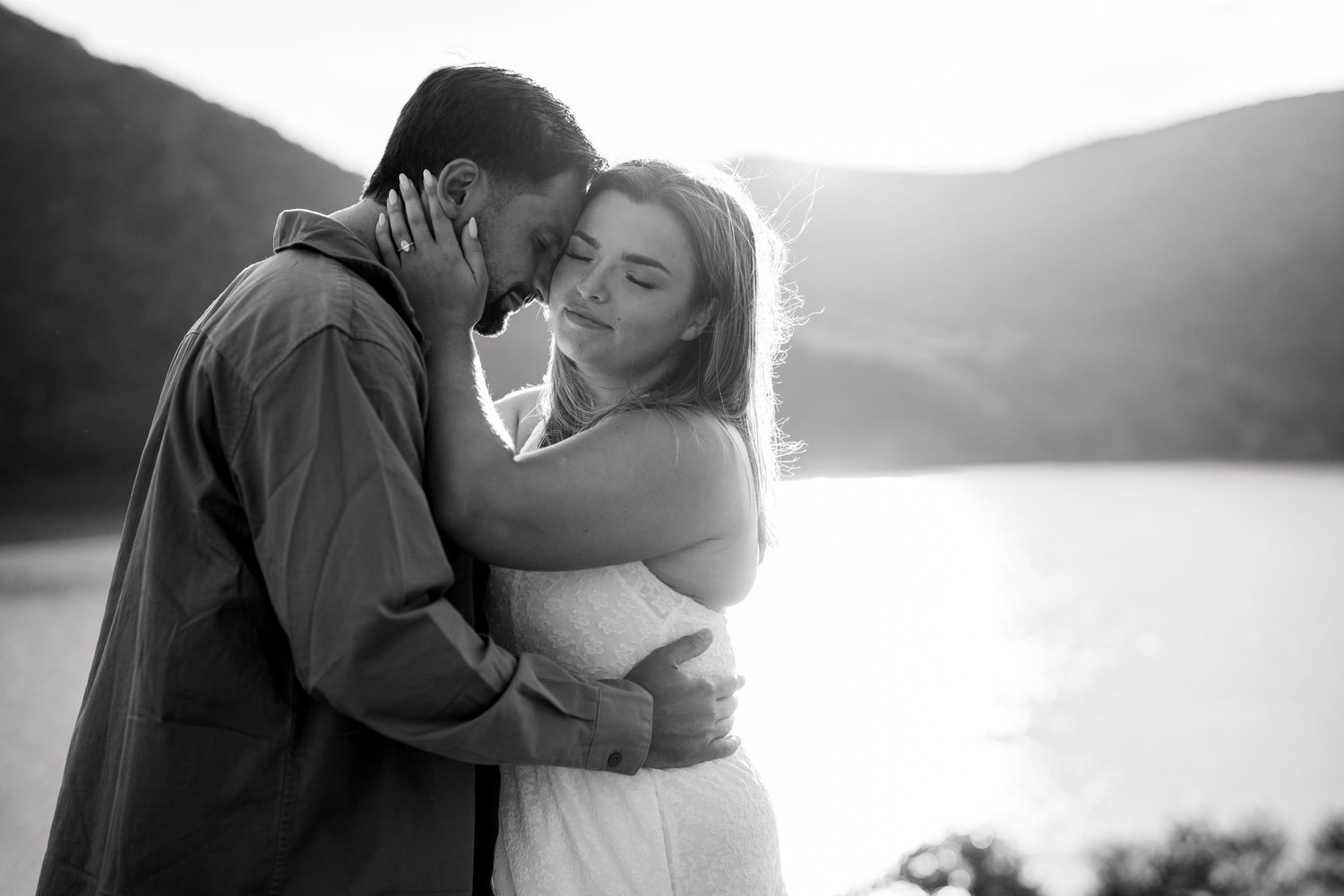 Black and white photo of a romantic couple embracing near a lake with mountain backdrop at sunset.