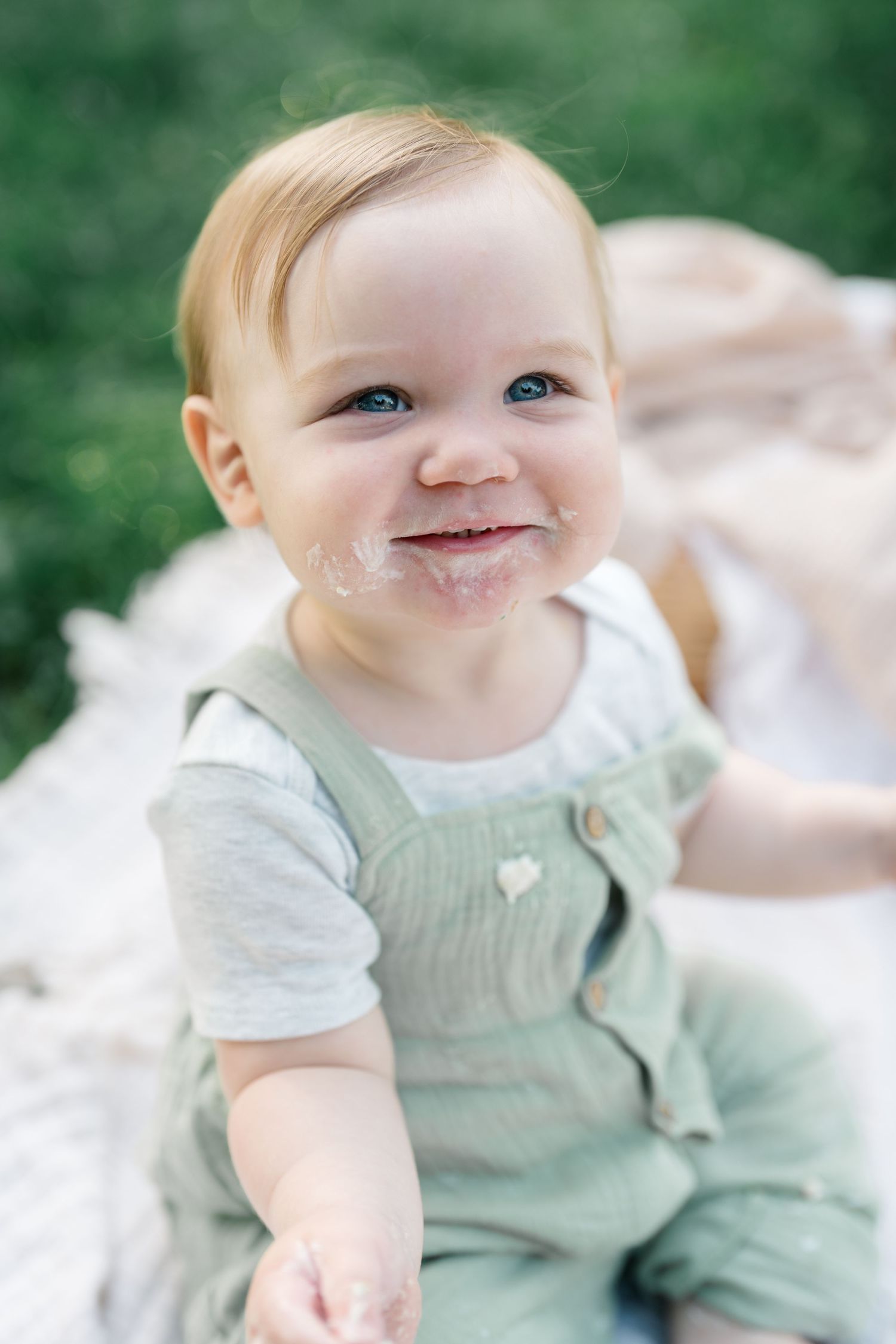 A cheerful baby with blue eyes and frosting on face sits on a blanket in a sage green romper outdoors.