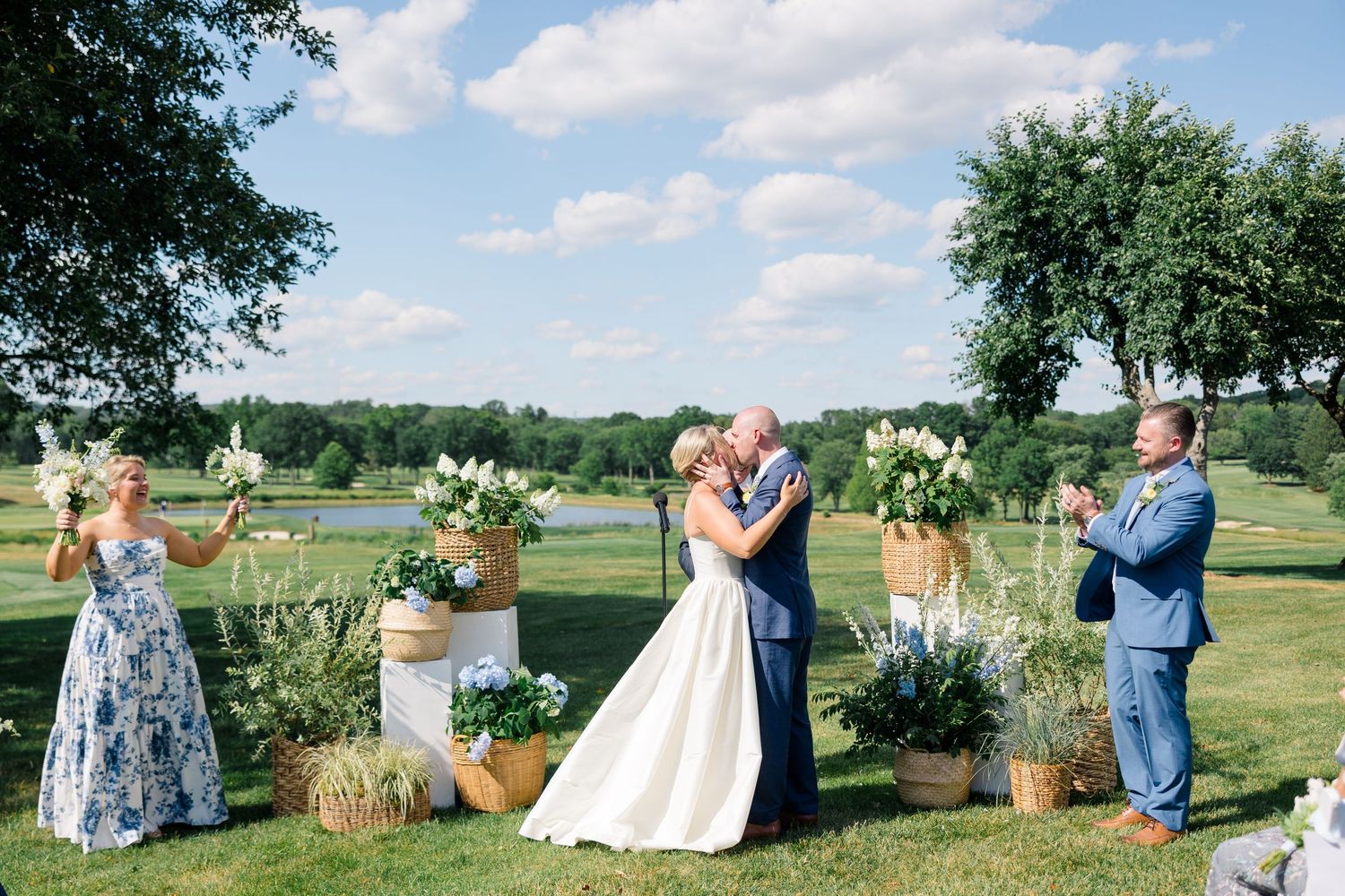 Newlyweds share their first kiss at an outdoor ceremony with floral arrangements and a scenic pond backdrop.