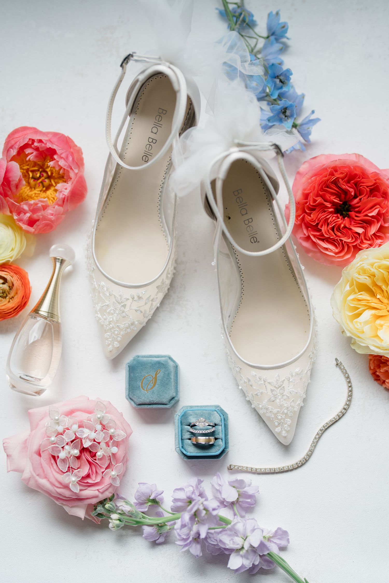 Elegant white lace bridal shoes styled with colorful flowers, wedding rings in blue velvet box, and delicate floral accents.