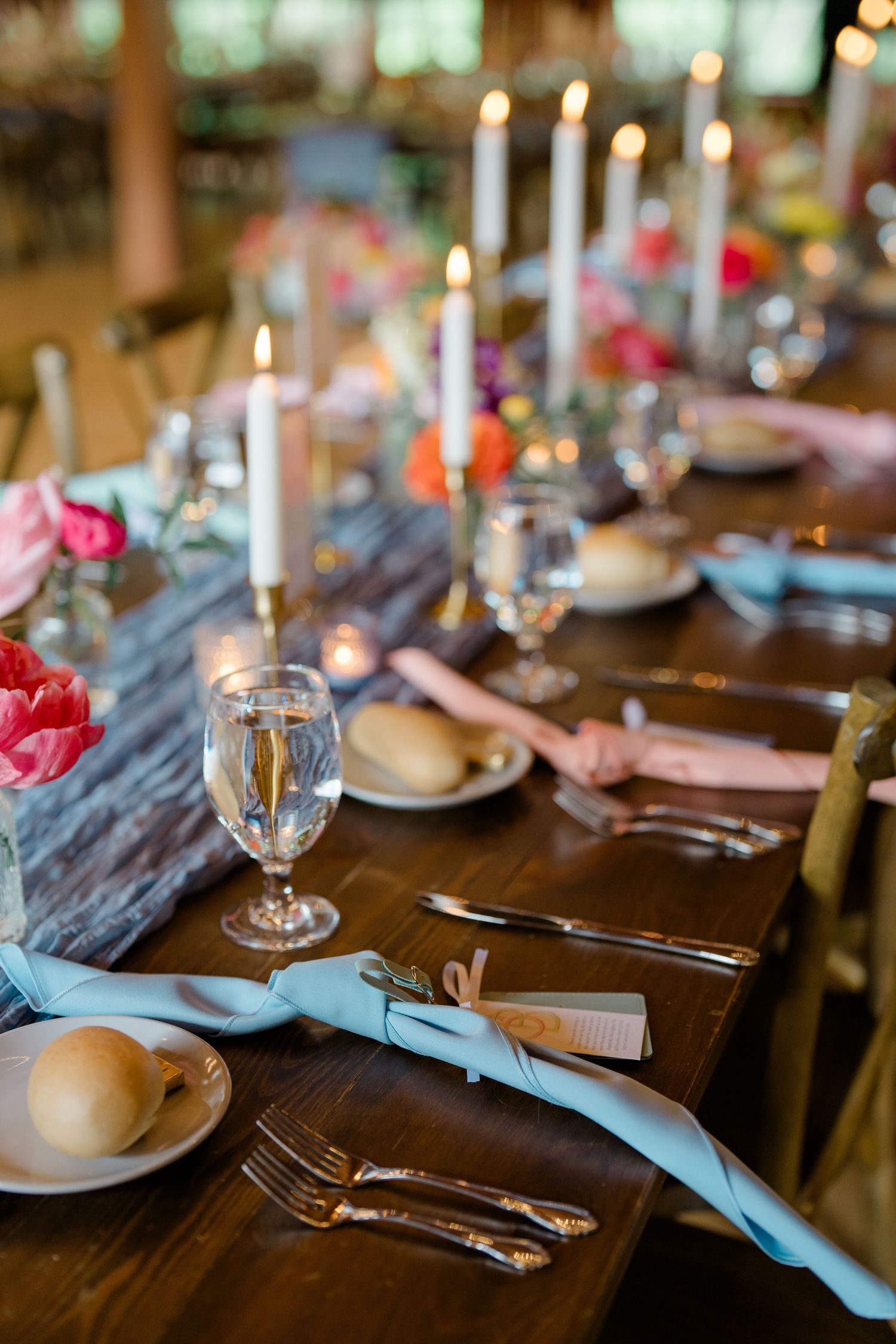 Elegant wedding reception table setting with pink accents, candles and blue napkins on rustic wood.
